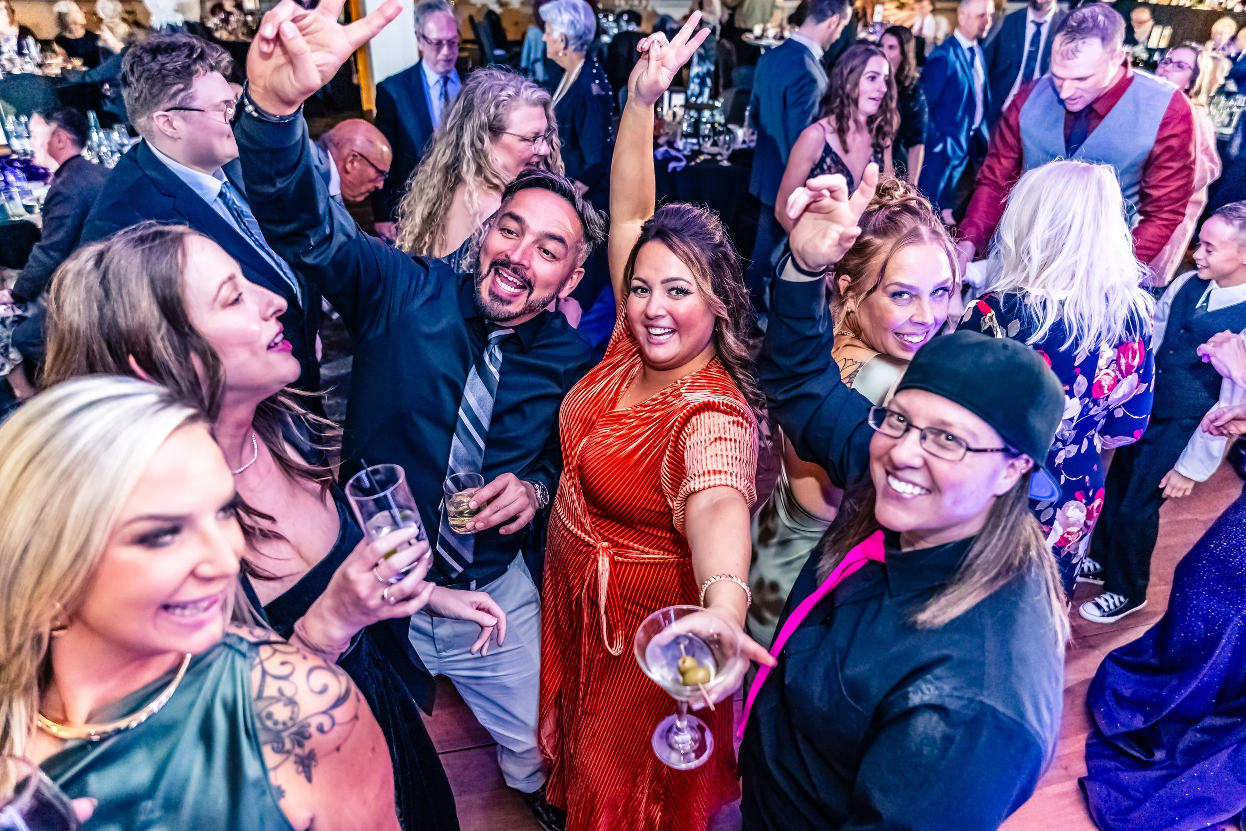 People dancing and celebrating at a party or wedding reception, with some holding drinks, and everyone smiling and showing lively expressions.