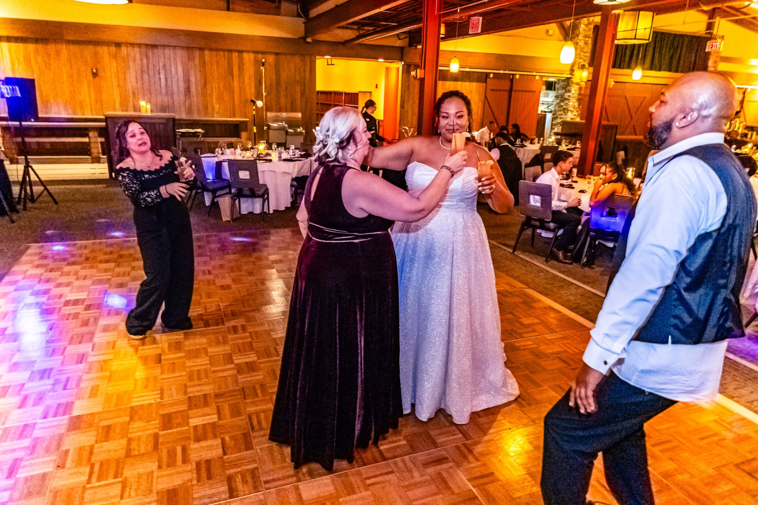 People dancing and socializing at a wedding reception with tables set for a meal in the background.