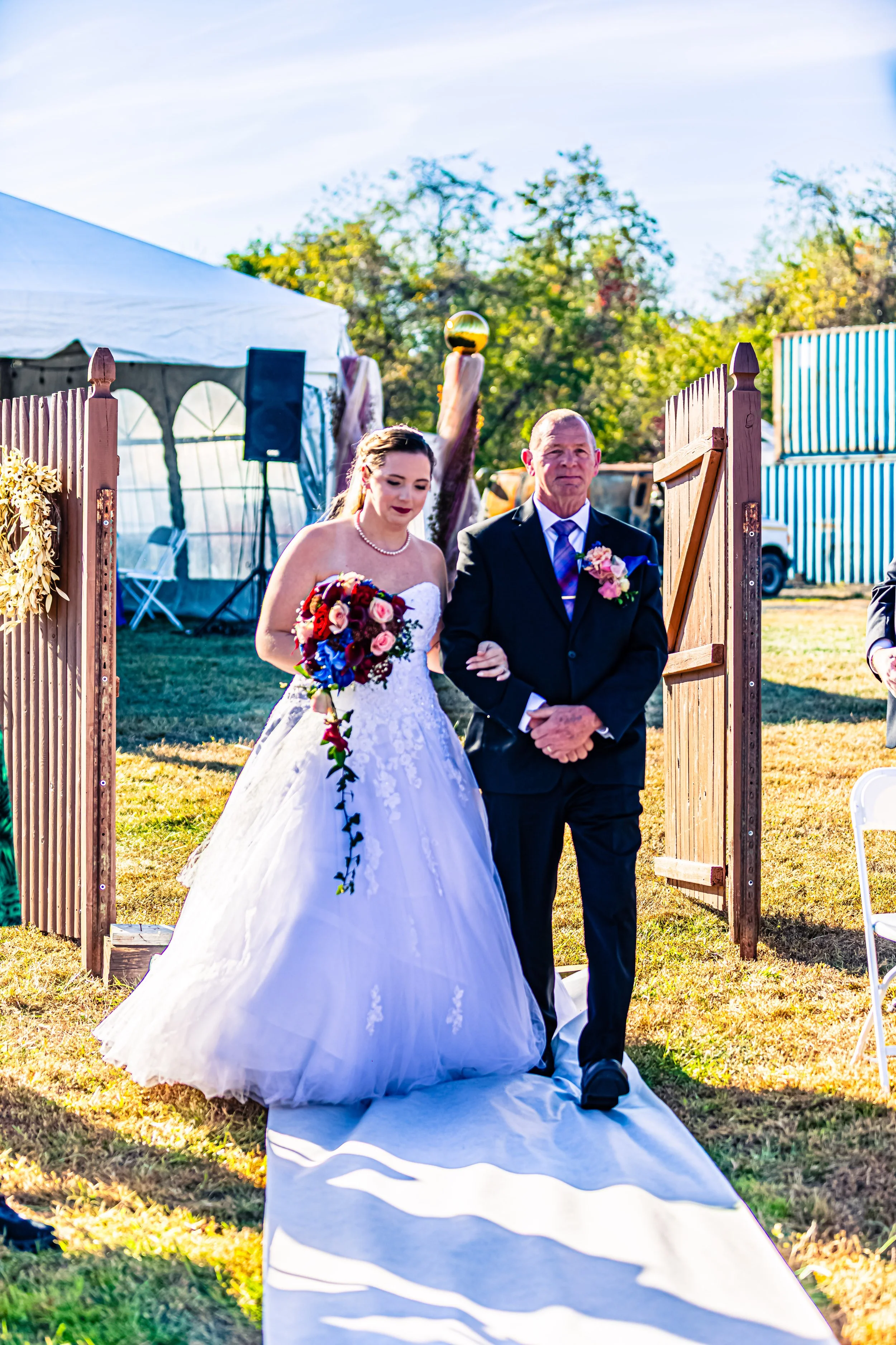 A bride dressed in a white wedding gown, holding a bouquet, walking down an outdoor aisle escorted by a man in a black suit with a boutonniere, during a wedding ceremony in a fenced yard with a tent and trees in the background.