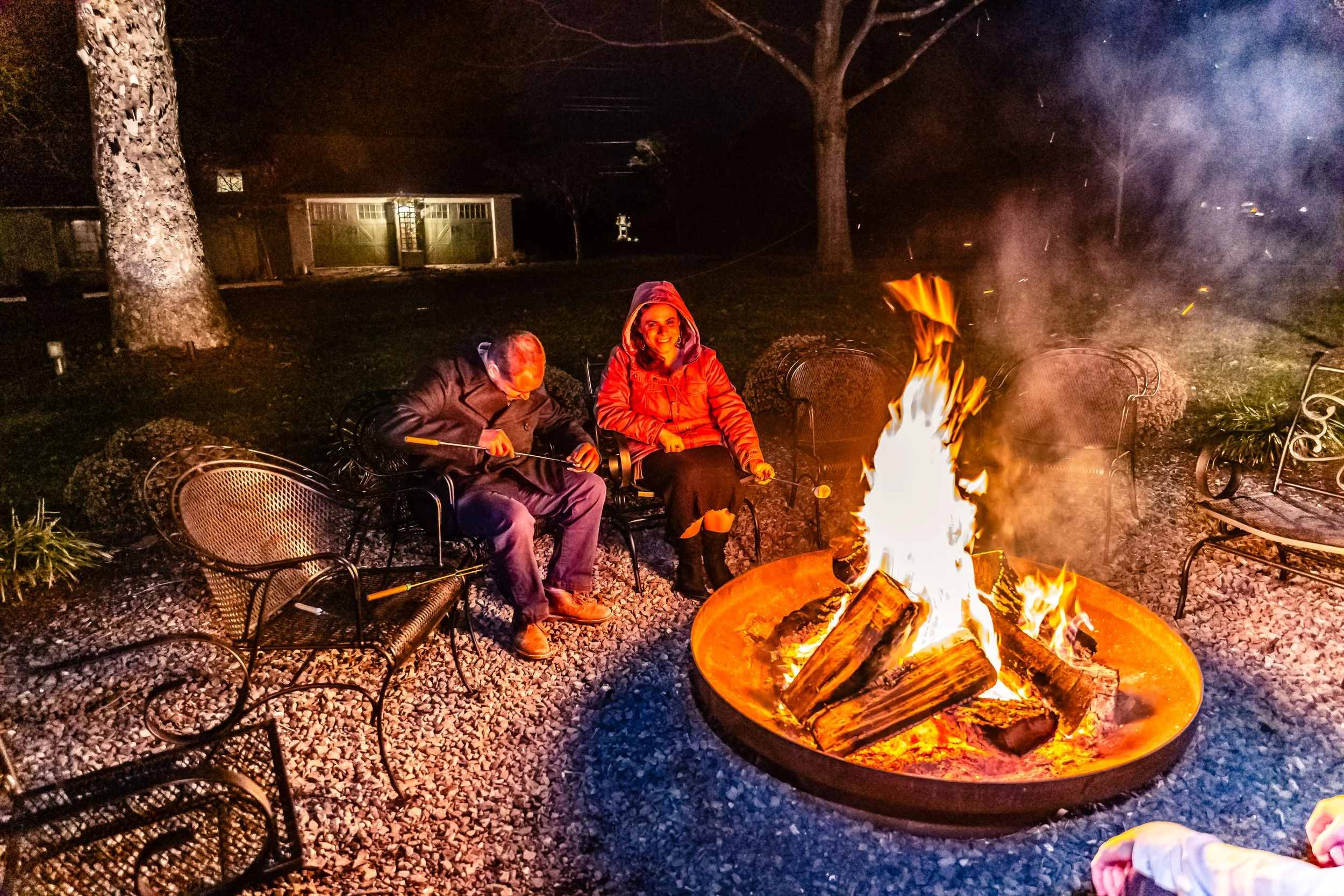 Two people sitting around a campfire at night outdoors, one man and one woman, with empty chairs around them.