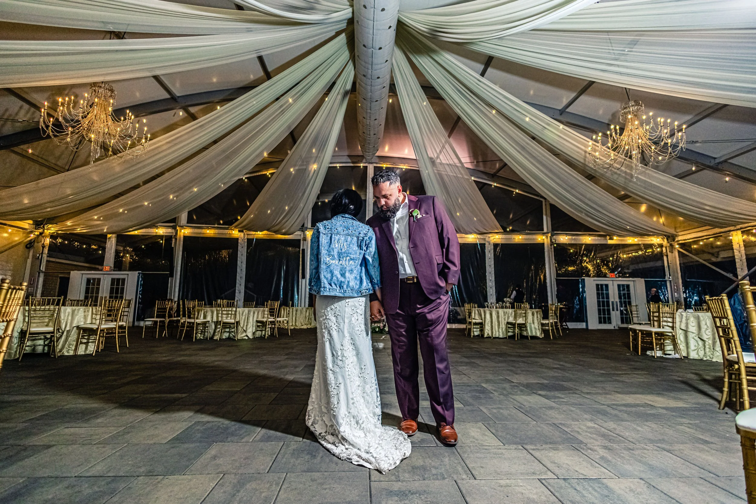 A bride and groom holding hands and standing close together in a decorated wedding reception tent with draped fabric and chandeliers, empty tables and chairs in the background.