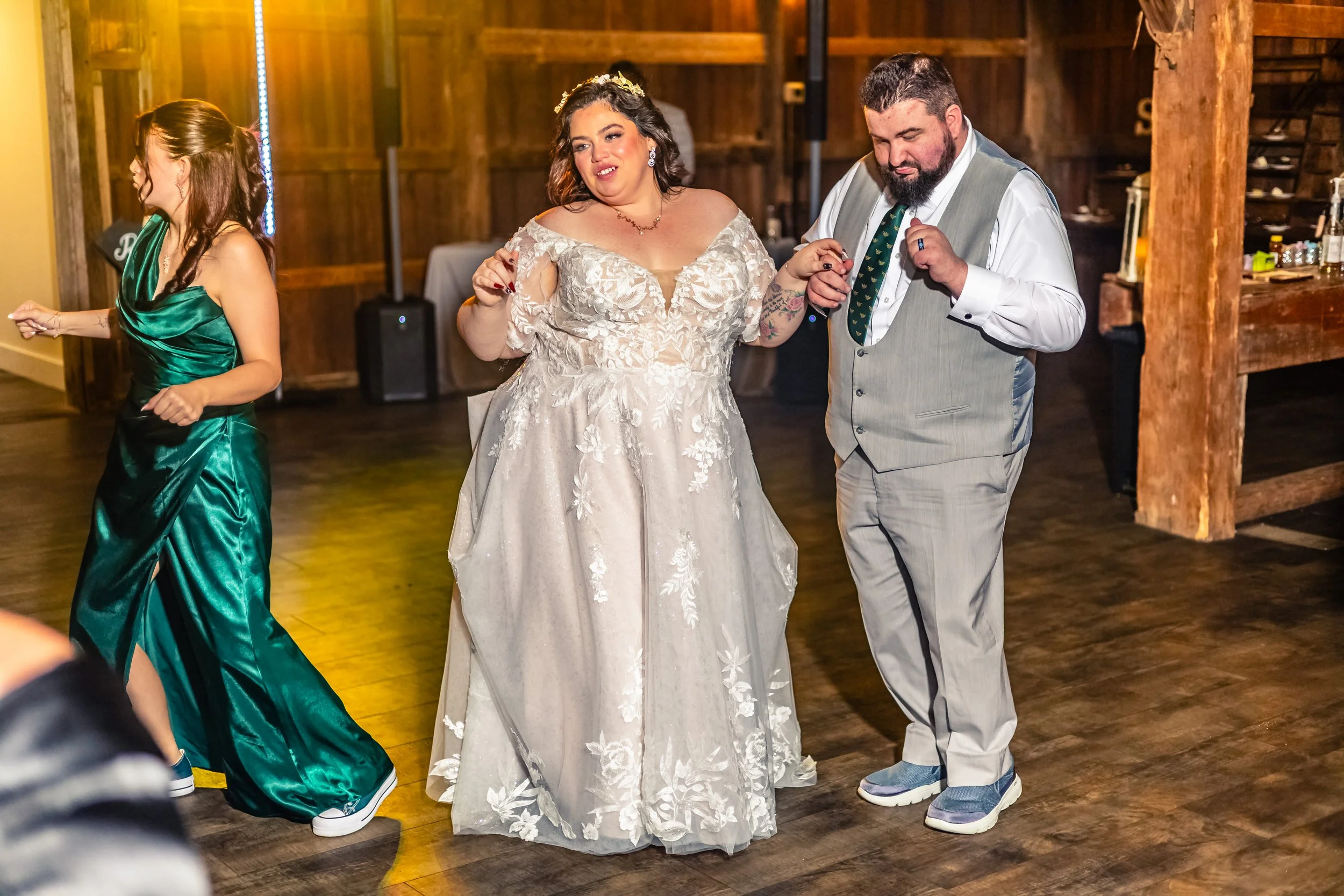 Bride and groom dancing together at a wedding reception, with a woman in a green dress dancing nearby in a rustic indoor venue with wooden walls and flooring.