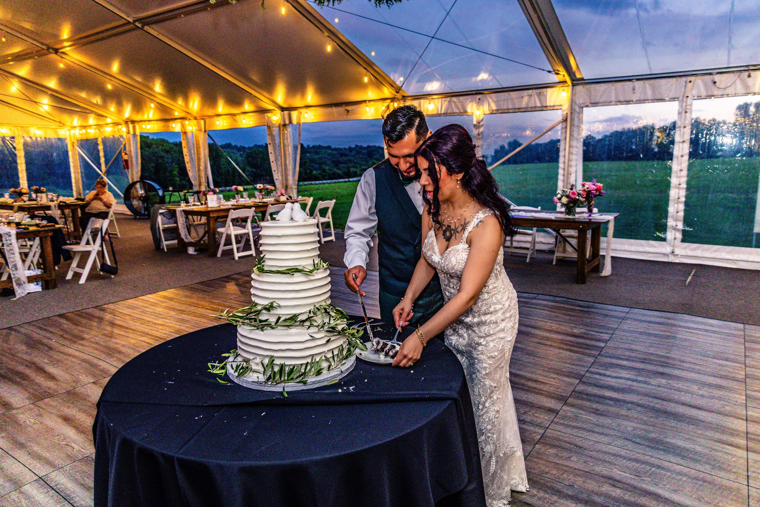A bride and groom cutting a wedding cake outside under a tent at dusk, with golden string lights overhead and a scenic green landscape in the background.