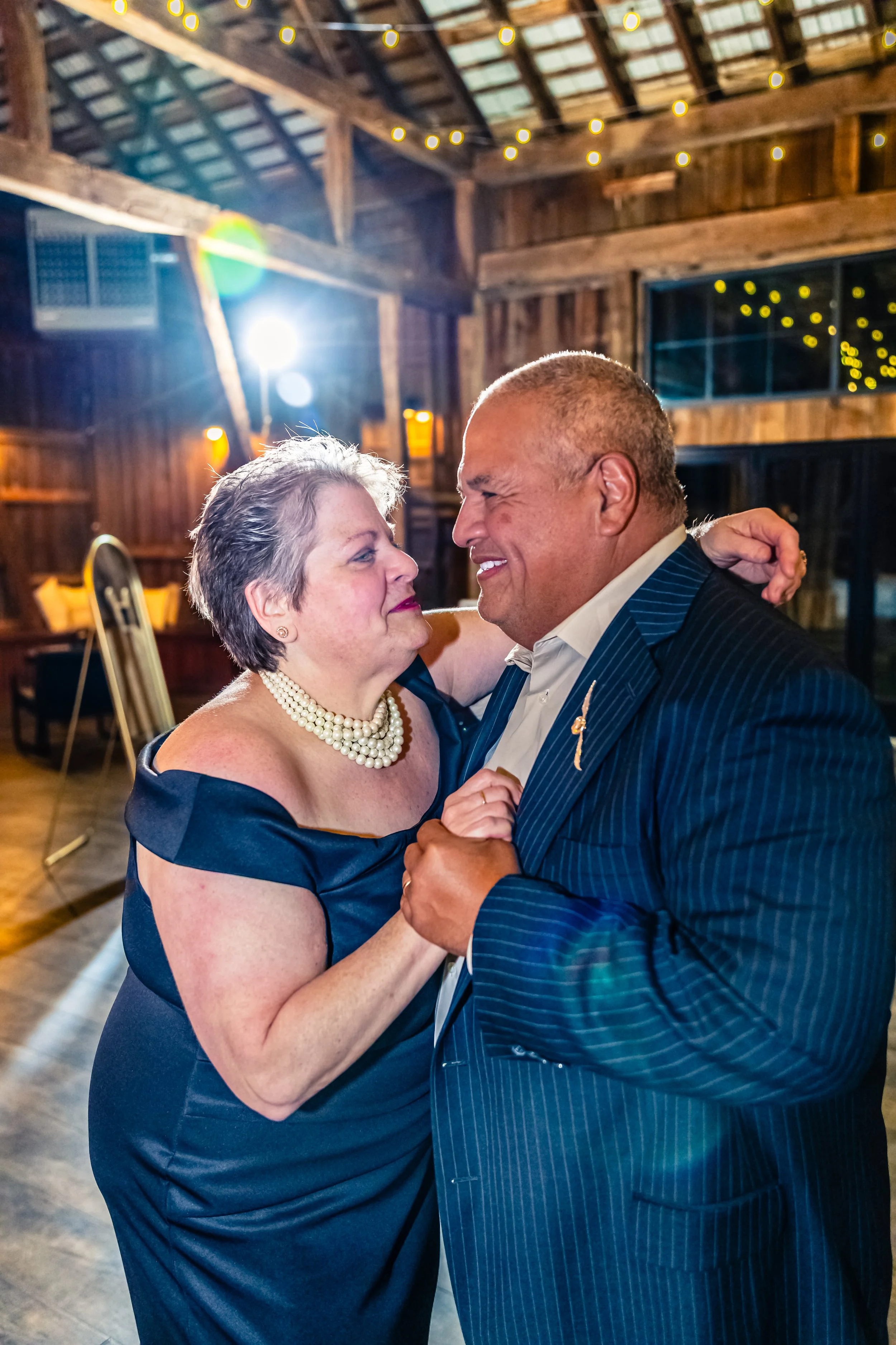 A senior woman in an off-shoulder navy dress and pearl necklace dancing with a senior man in a pinstripe suit at a rustic indoor event, with string lights overhead.
