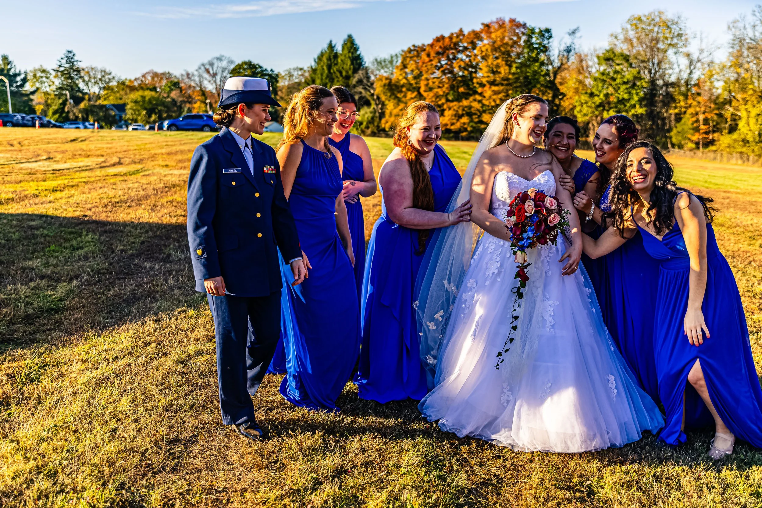 A group of women, including a bride in a white wedding gown holding a bouquet and wearing a veil, and bridesmaids dressed in blue, standing outdoors on a grassy field during sunset, smiling and interacting.