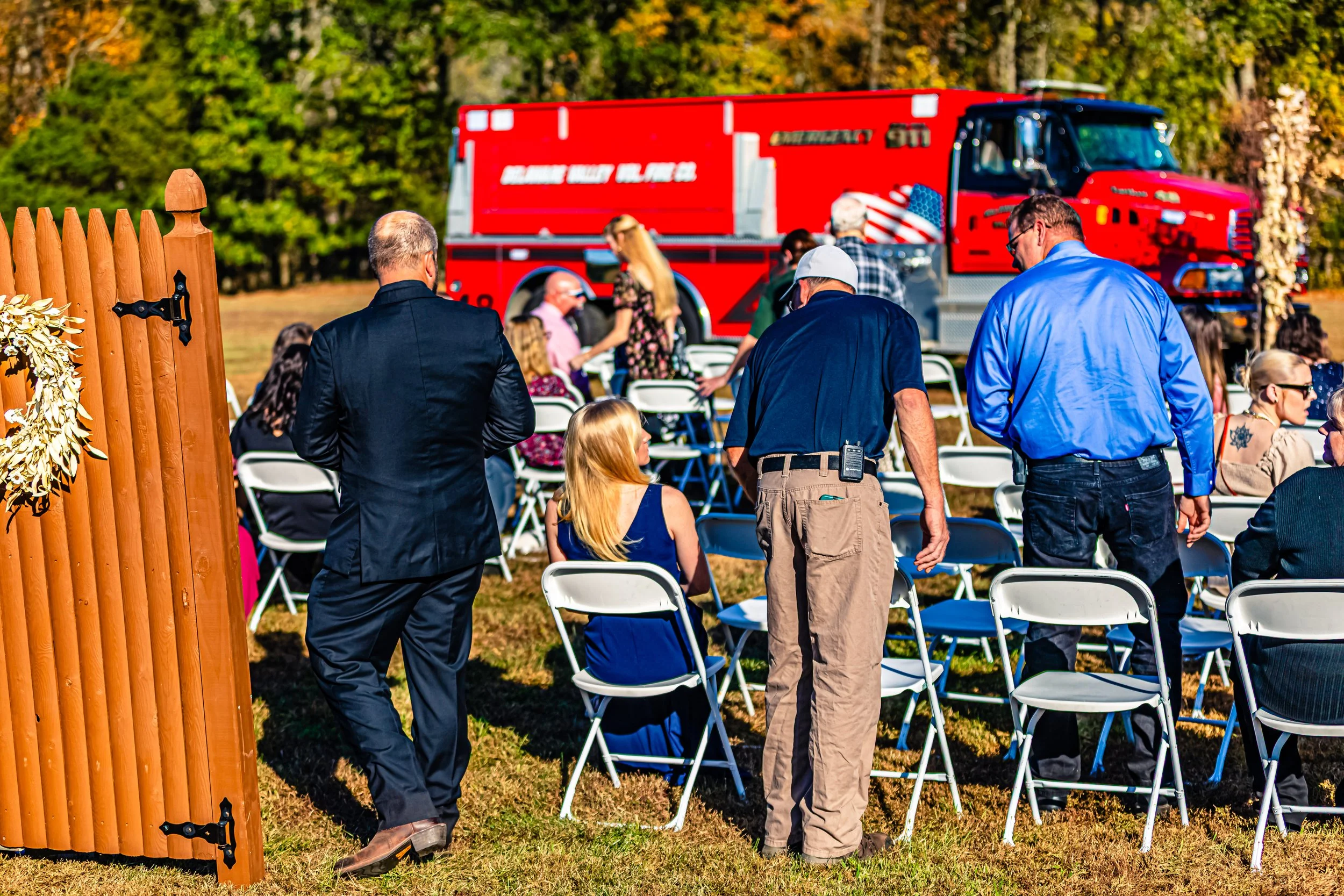 People attending a memorial or outdoor event with a red fire truck in the background, some seated and some standing on a grassy area surrounded by trees.