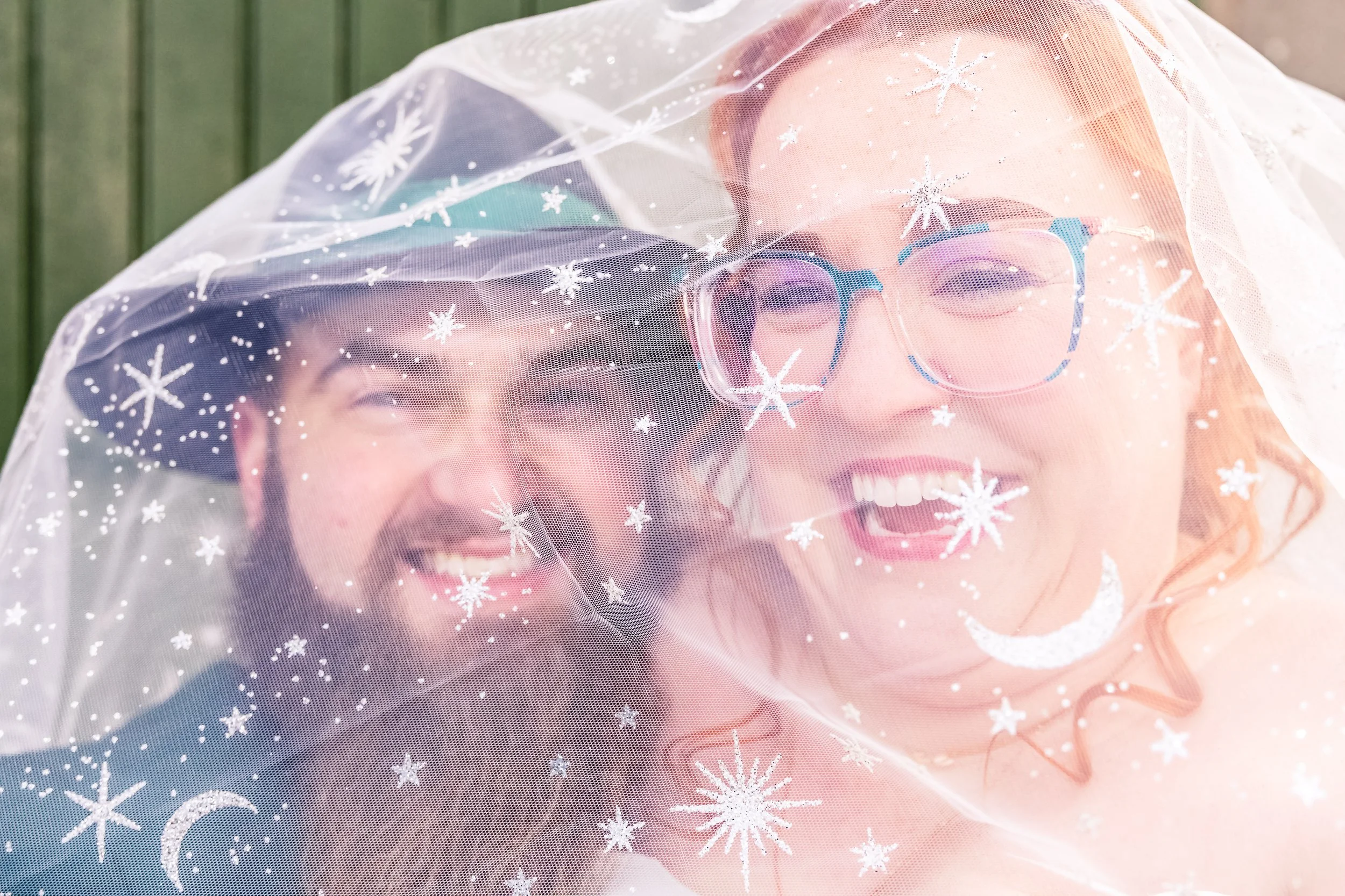 A smiling man and woman under a sheer veil with snowflake, moon, and star patterns, outdoors.