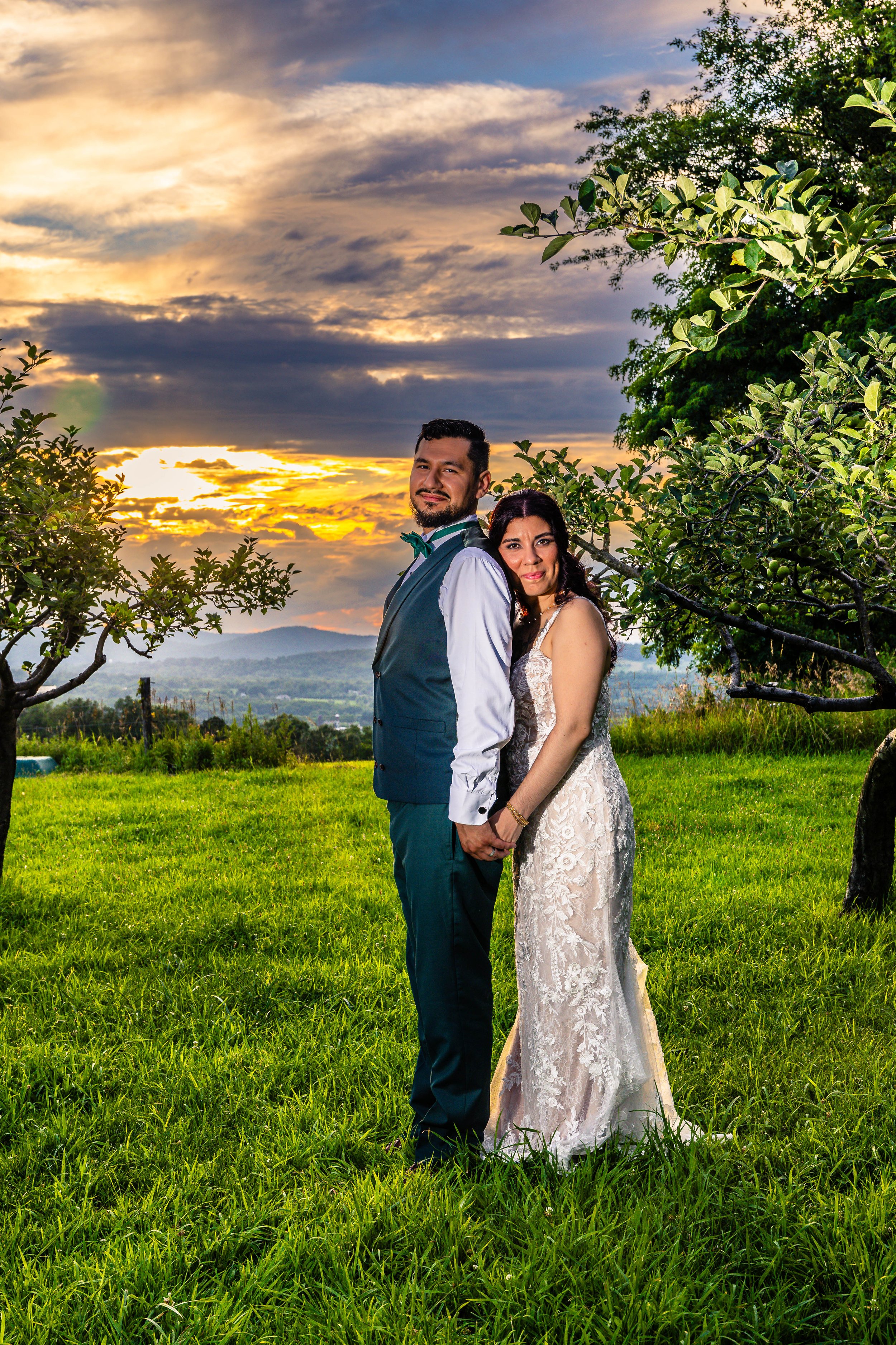 A bride and groom in wedding attire standing hand in hand outdoors during sunset, with trees and mountains in the background.