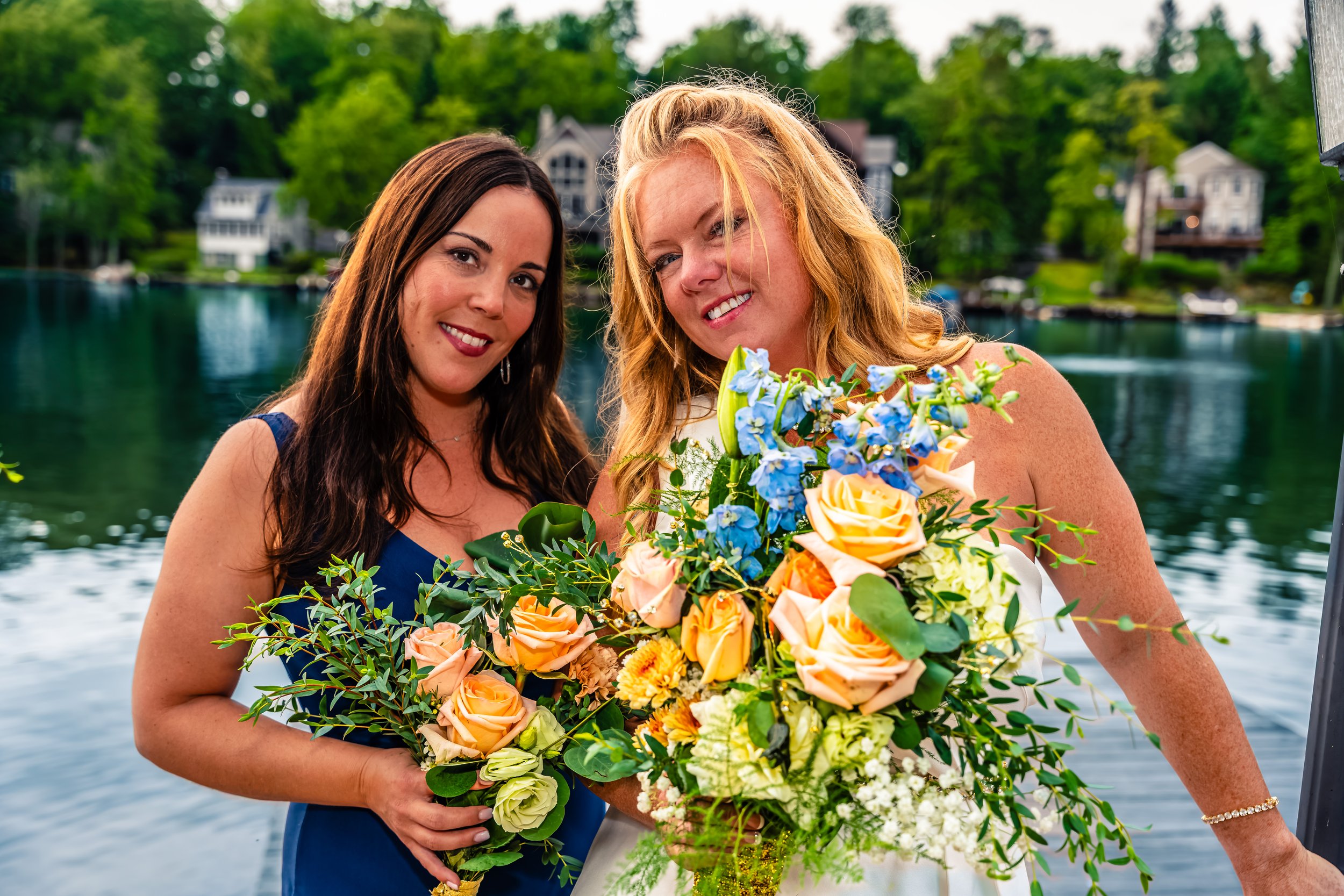 Two women holding large floral bouquets by a lake with houses and trees in the background.