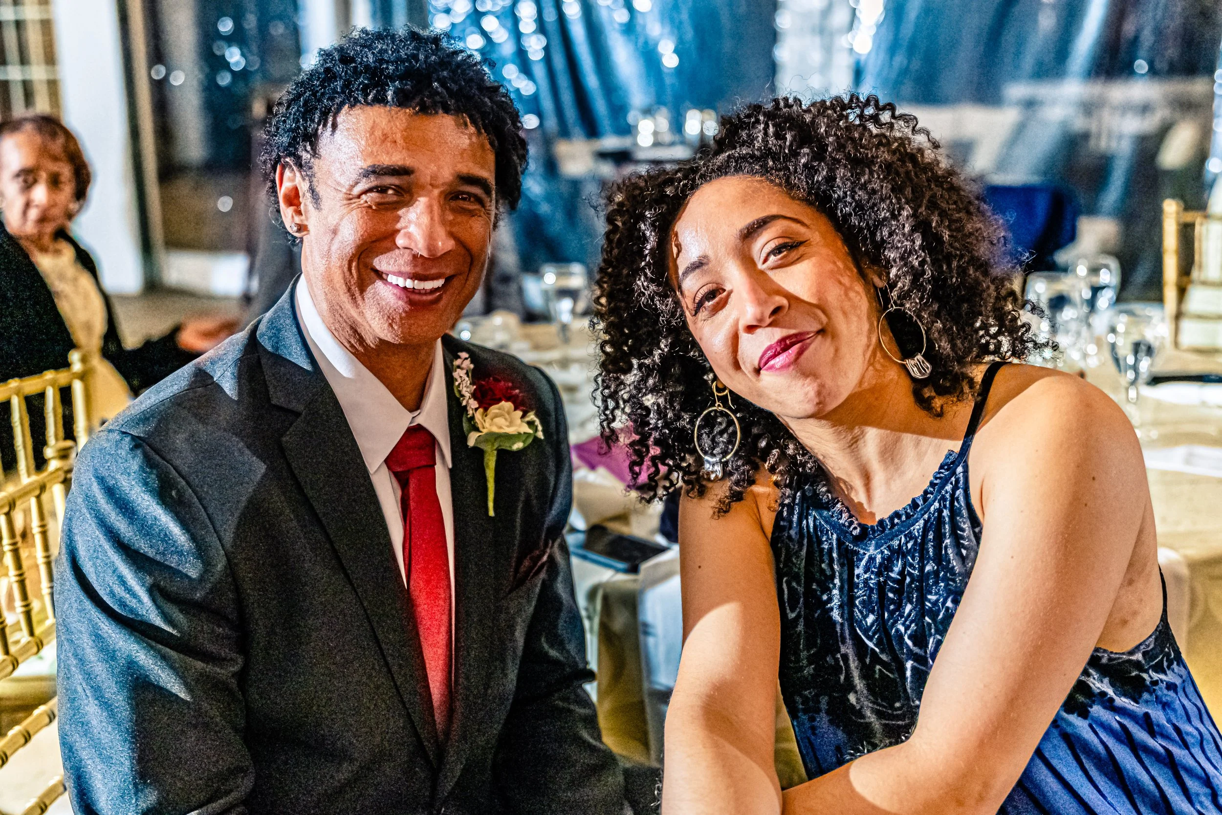 A smiling couple dressed in formal attire at a celebration event, with a woman in a blue dress with curly black hair and a man in a dark suit with a red tie and a boutonniere, sitting at a decorated table with other guests and banquet settings in the