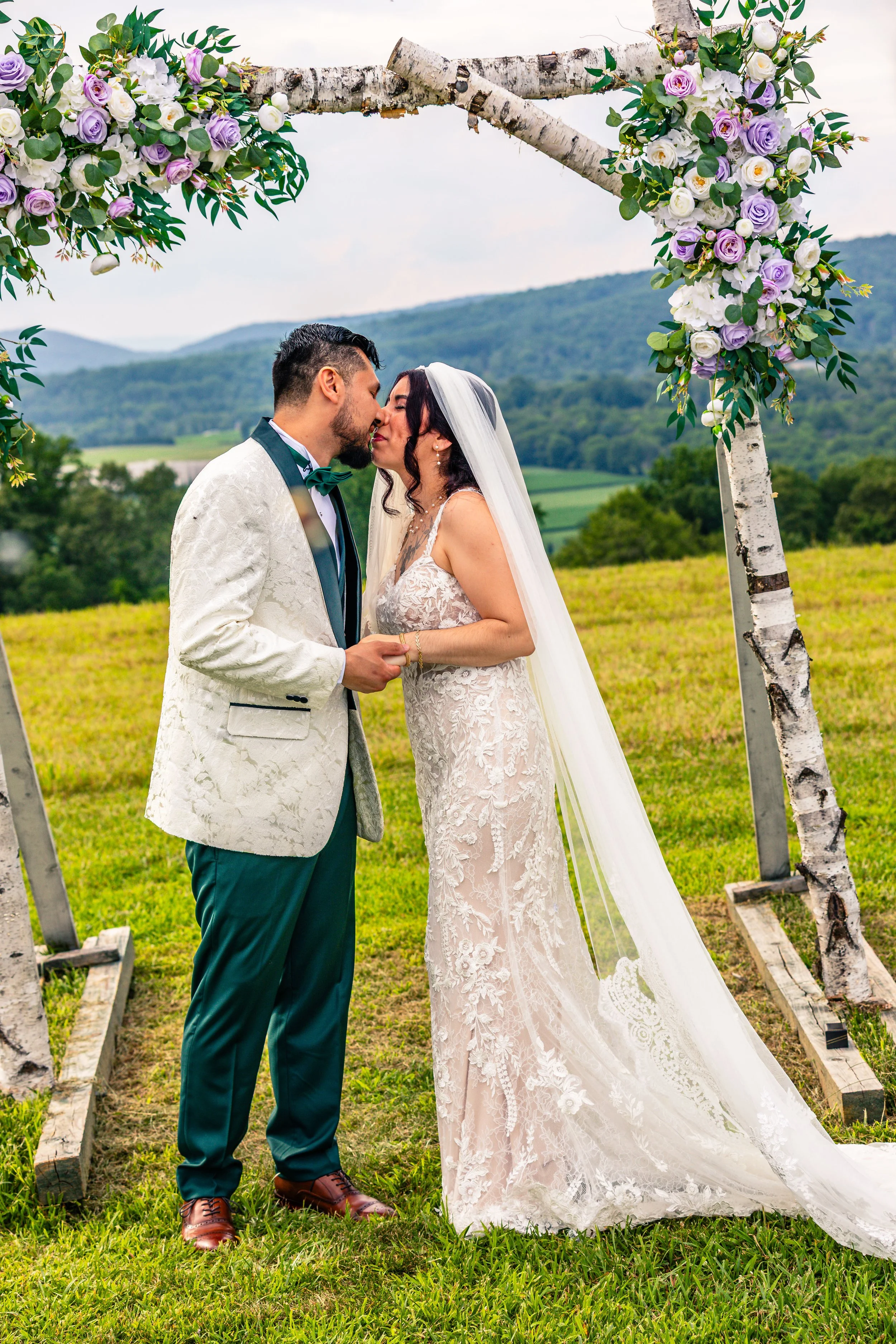 A bride and groom share a kiss during their outdoor wedding ceremony under a rustic arch decorated with white and purple flowers, set against a scenic landscape of green hills and mountains.