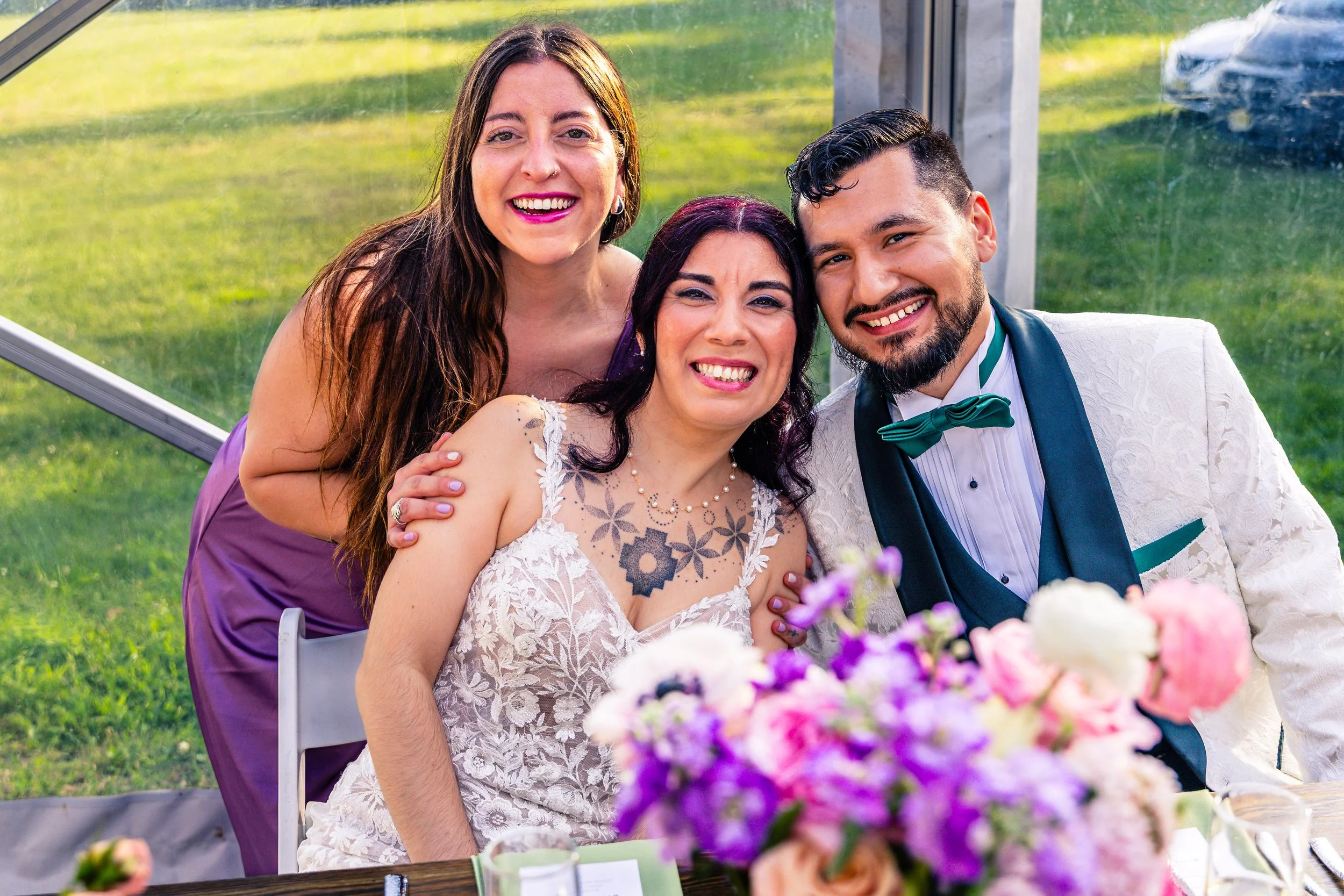 Four people smiling at a wedding reception, with two women and a man in formal attire and a bride with tattoos in a wedding dress, all sitting at a table with a colorful flower centerpiece outside.