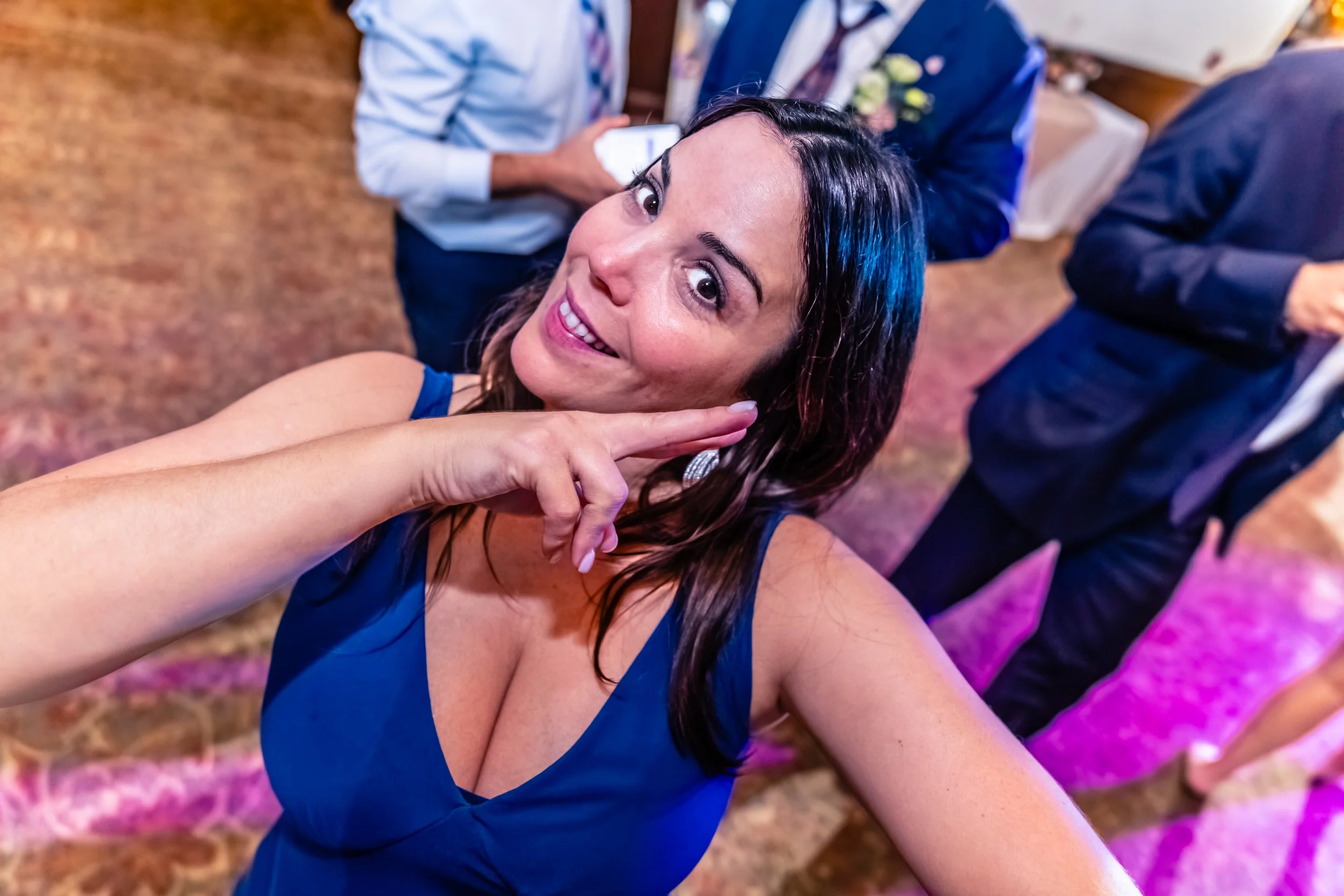 Woman in blue dress taking a selfie at a social event with people in the background.