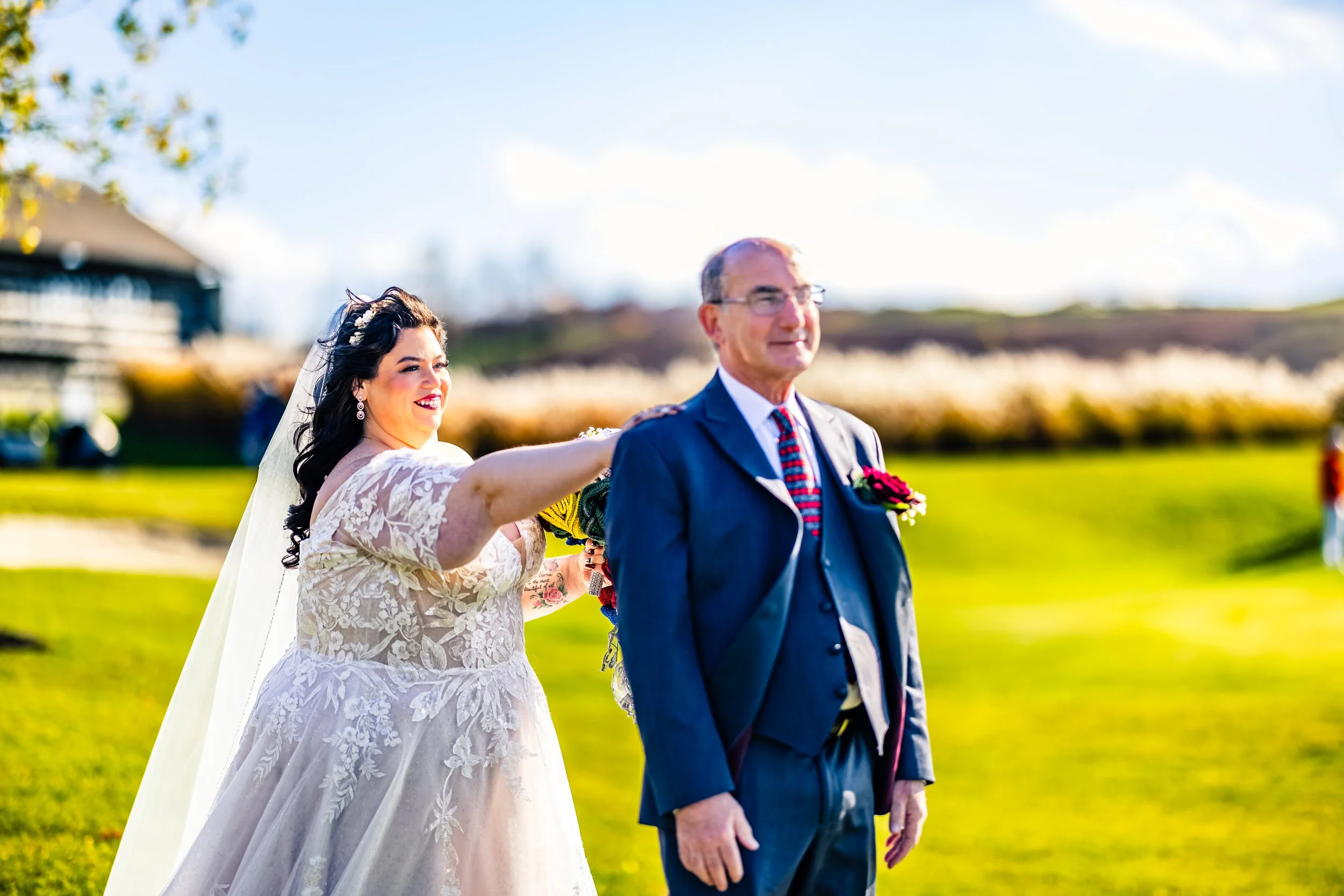 A bride is holding a bouquet and appears to be playfully pulling on the groom's tie during an outdoor wedding ceremony on a sunny day.
