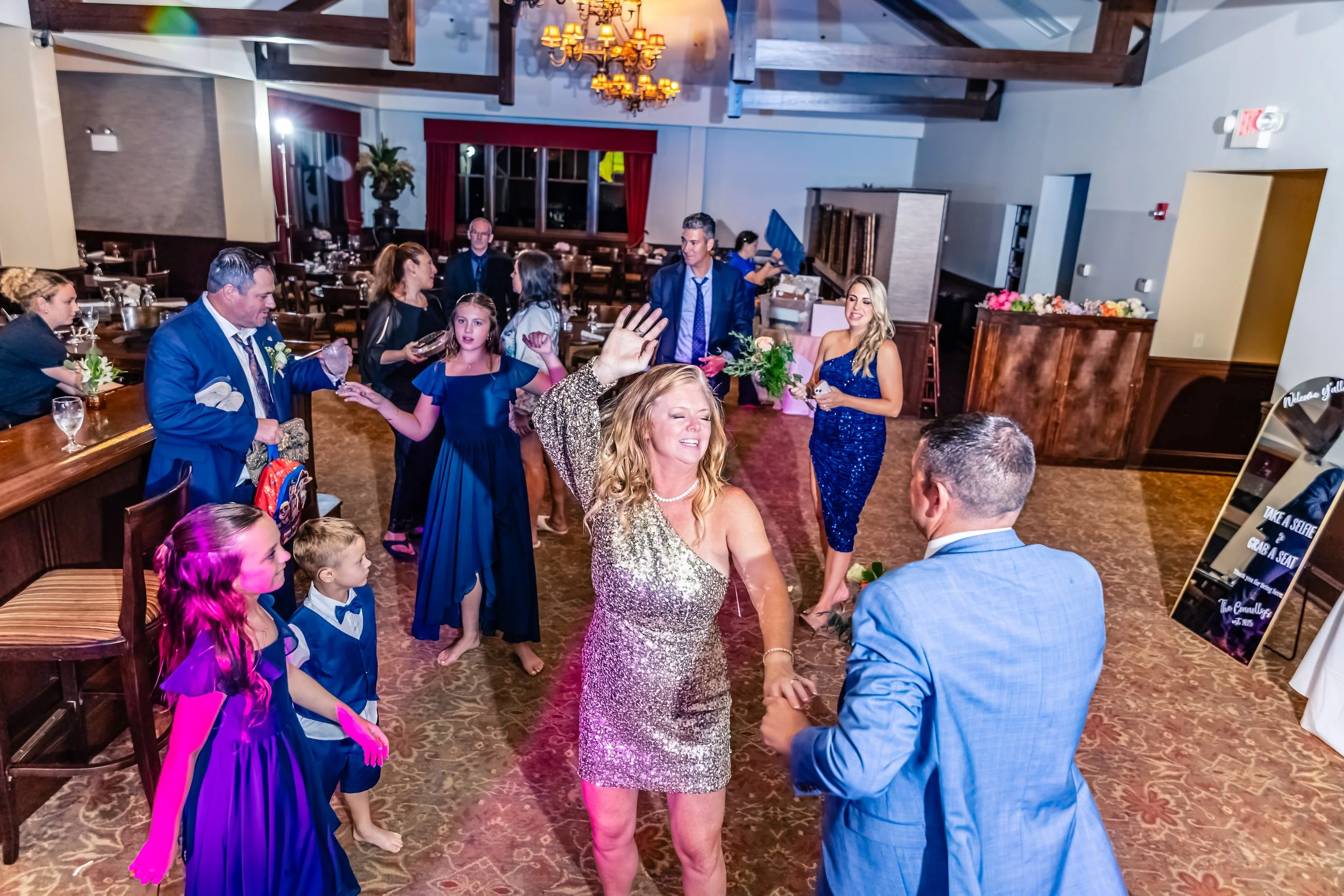People dancing and celebrating at a wedding reception, including a woman in a sparkly dress holding hands with a man in a blue suit, surrounded by children and other guests in a decorated banquet hall.