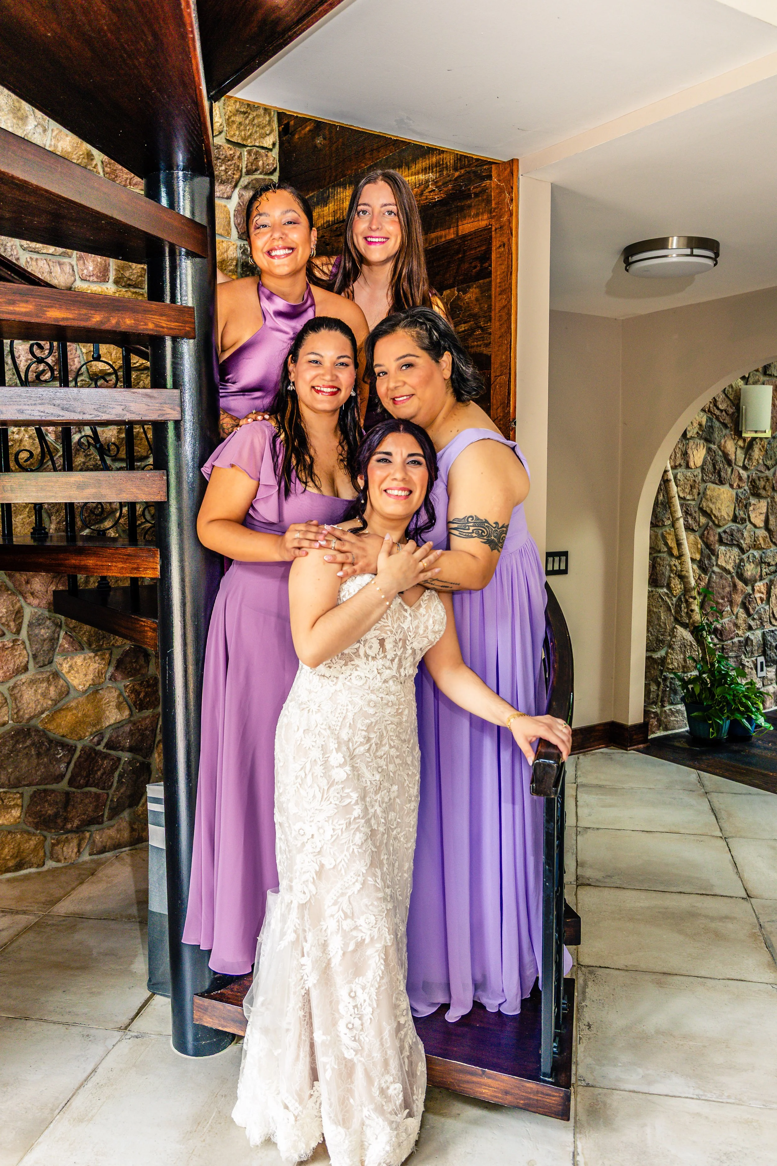 Six women, with one in a wedding gown and five in lavender dresses, posing together inside a house near a wooden staircase and a stone wall.