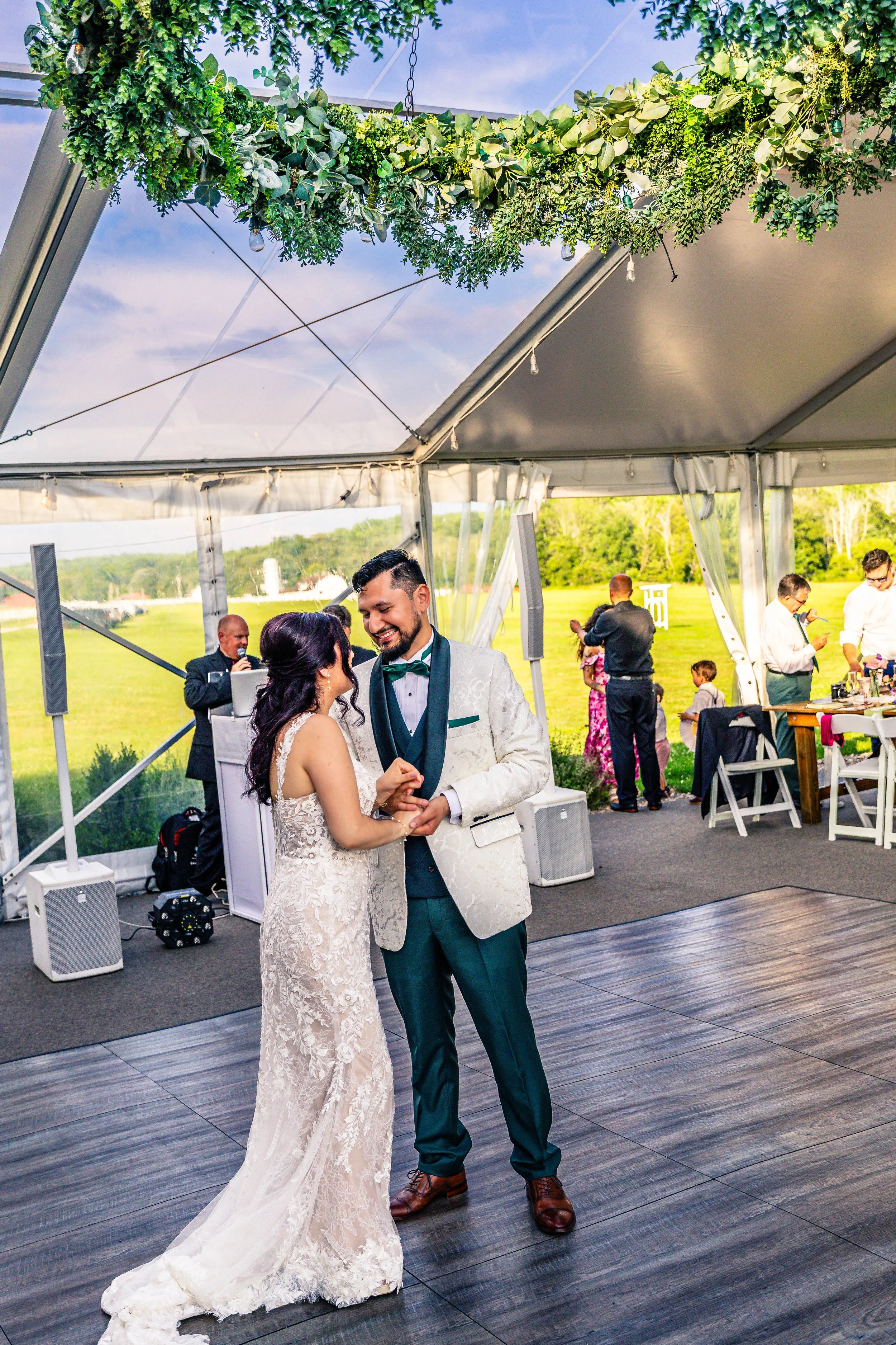 A bride and groom dancing at their wedding reception inside a decorated tent, with a view of green fields and blue sky in the background.