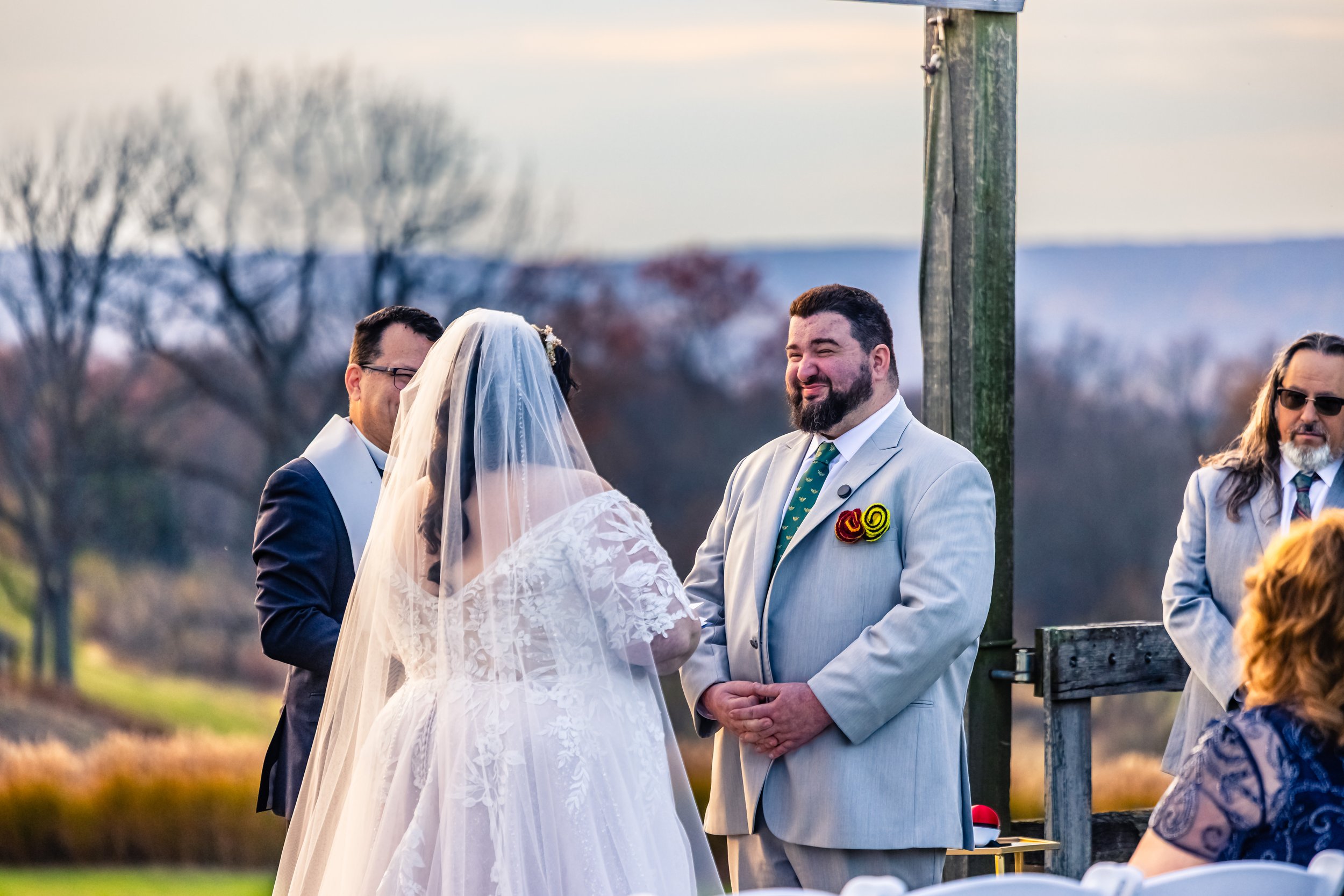 A wedding ceremony outdoors with a bride and groom facing each other, with officiant and guests present. Bride wears a white lace wedding dress and veil, groom wears a light gray suit with colorful lapel pins, and the background shows a scenic landsc