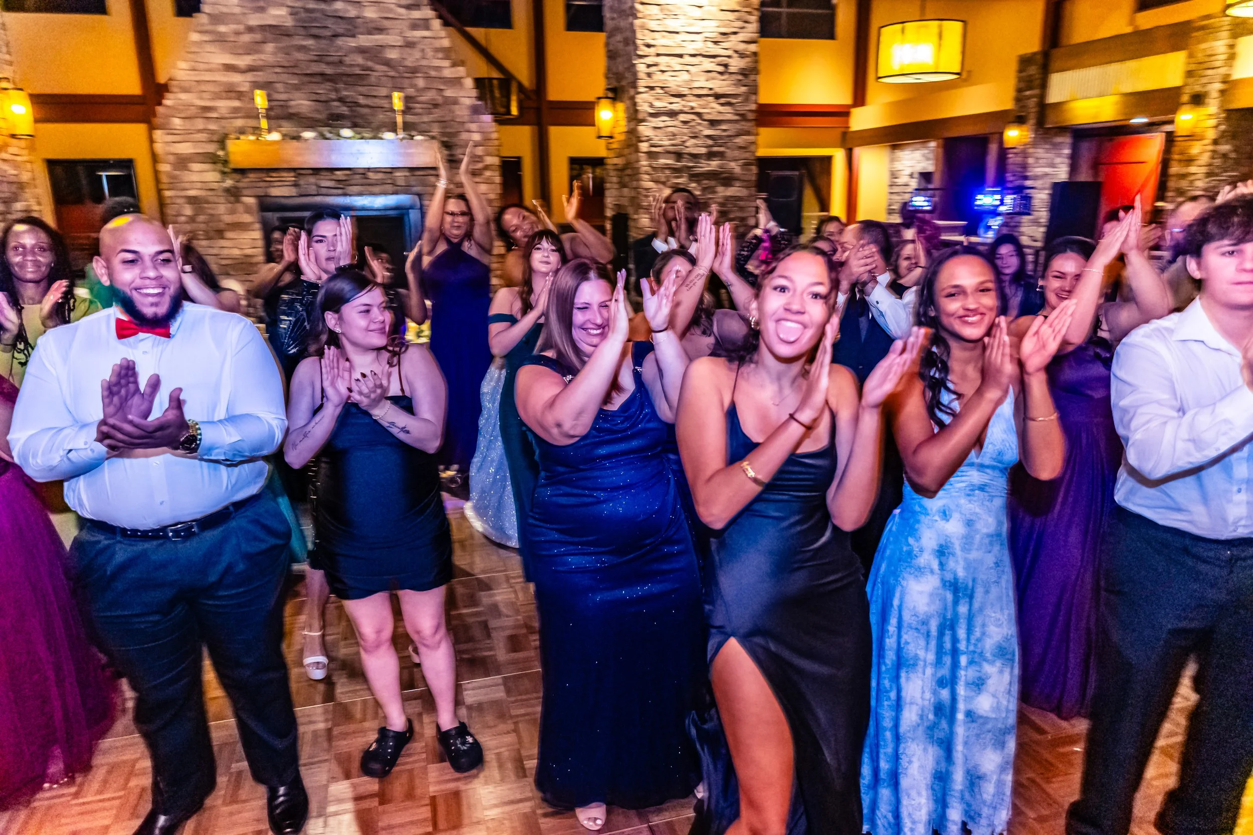 Group of people dancing and celebrating at a party or wedding reception in a decorated venue with stone fireplace and warm lighting.