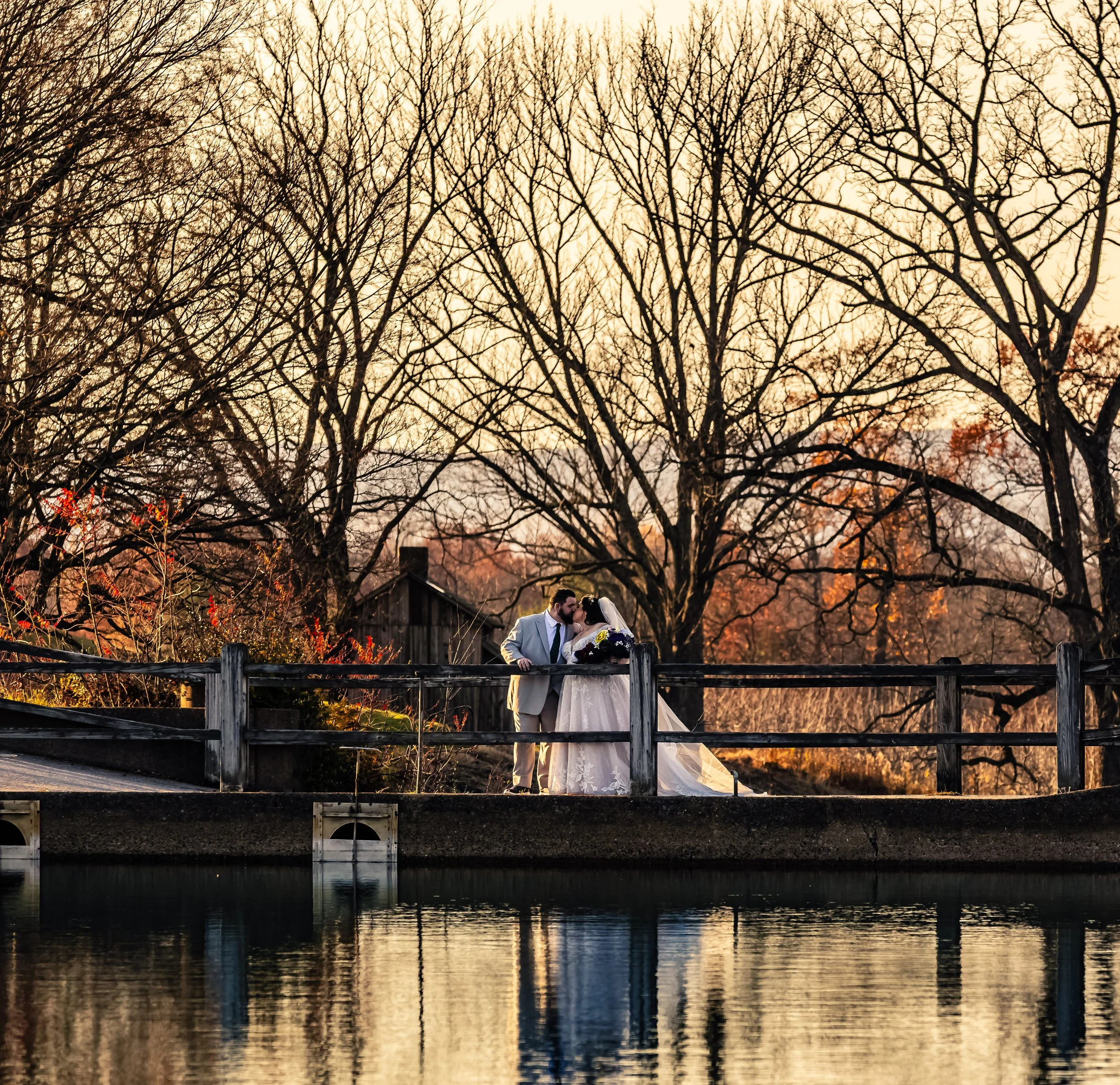 A bride and groom sharing a kiss on a bridge near a pond at sunset with bare trees in the background.