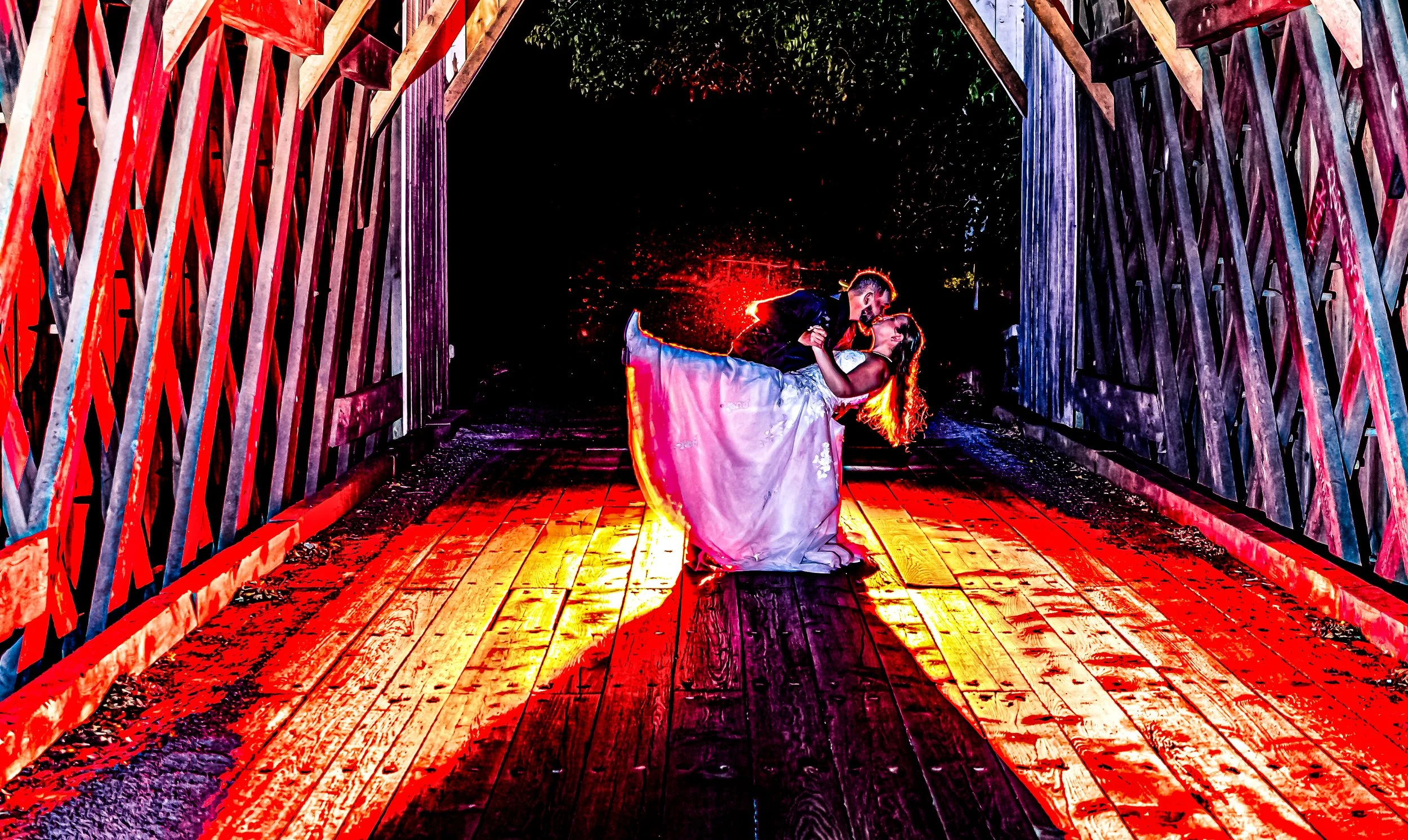A couple dancing closely on a wooden bridge at night, illuminated by colorful lighting that casts vibrant red and yellow shadows on the floor and surrounding structures.