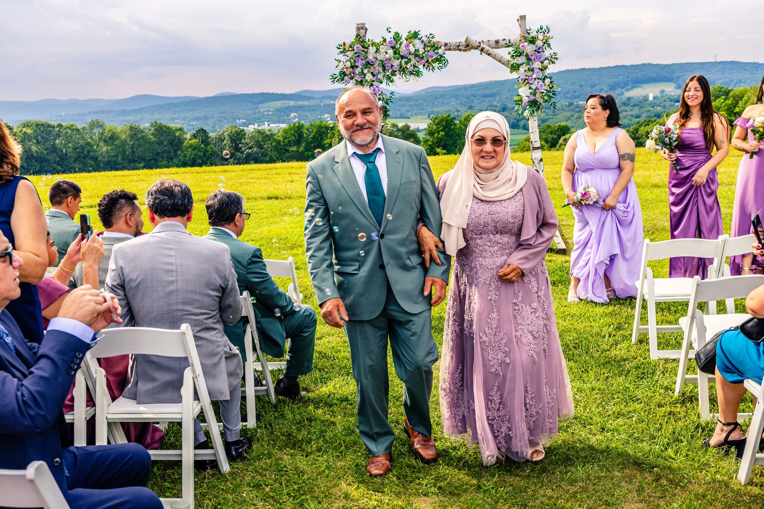 A couple walks down the aisle at an outdoor wedding ceremony with guests on either side. The man is in a teal suit and the woman is in a lavender lace dress with a light beige headscarf. Bridesmaids in lavender gowns stand behind them, holding bouque