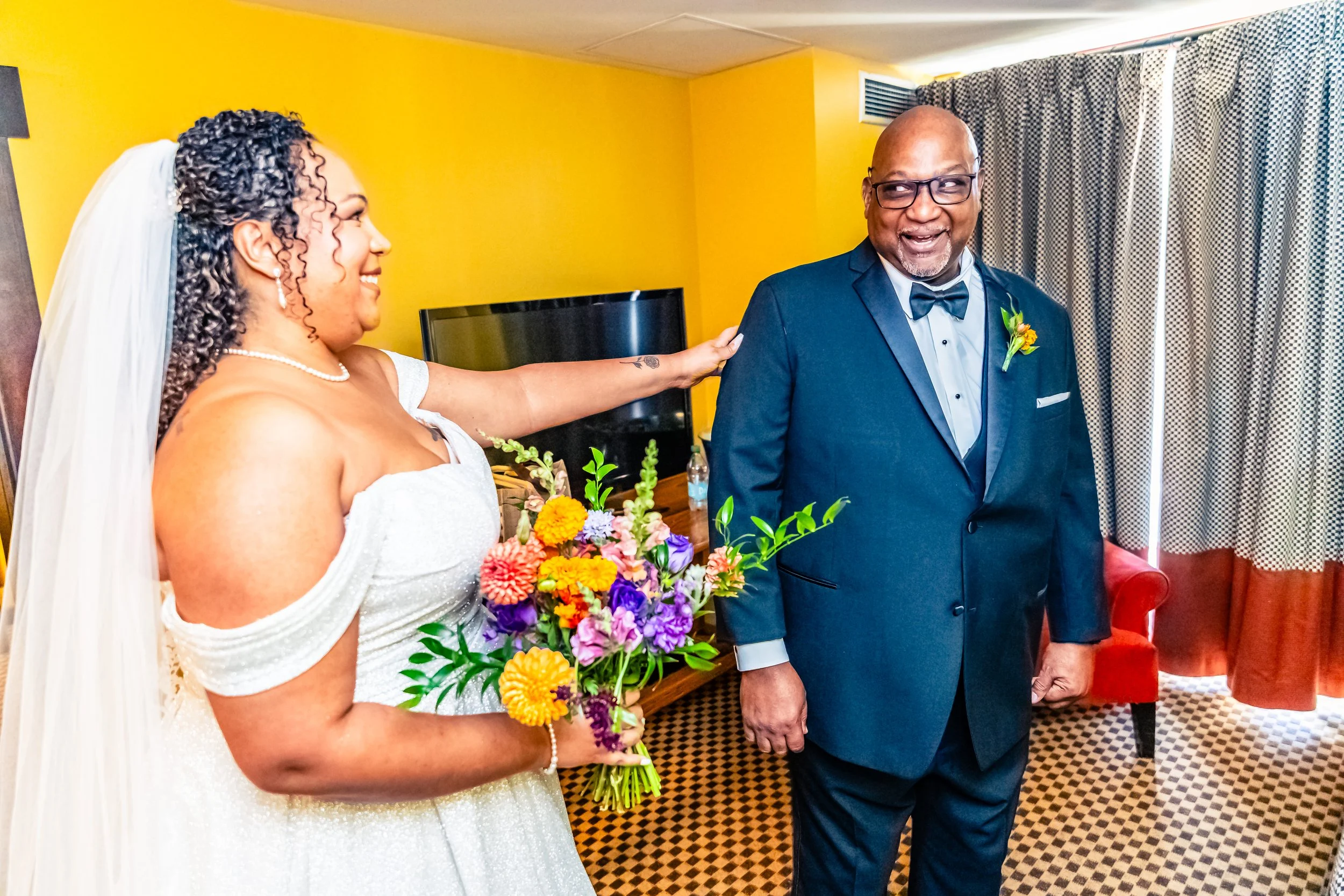 A bride in a white wedding dress holding a colorful bouquet extends her hand to touch the shoulder of a smiling, older man in a black tuxedo with a bow tie, in a warmly decorated room.