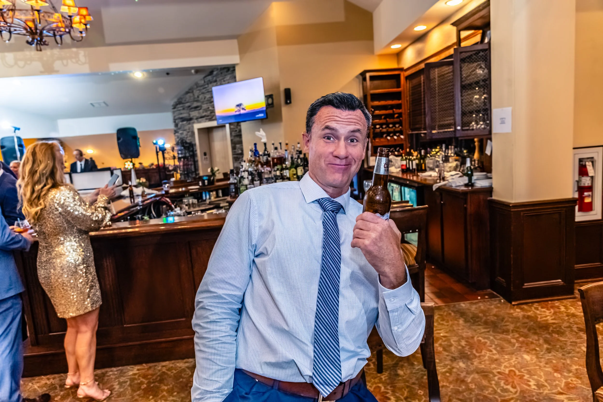 A man in a dress shirt and tie holding a beer bottle, smiling, at a bar. A woman in a gold sequined dress is in the background at the bar, looking at her phone. The setting appears to be a lively indoor bar or restaurant with warm lighting and wooden