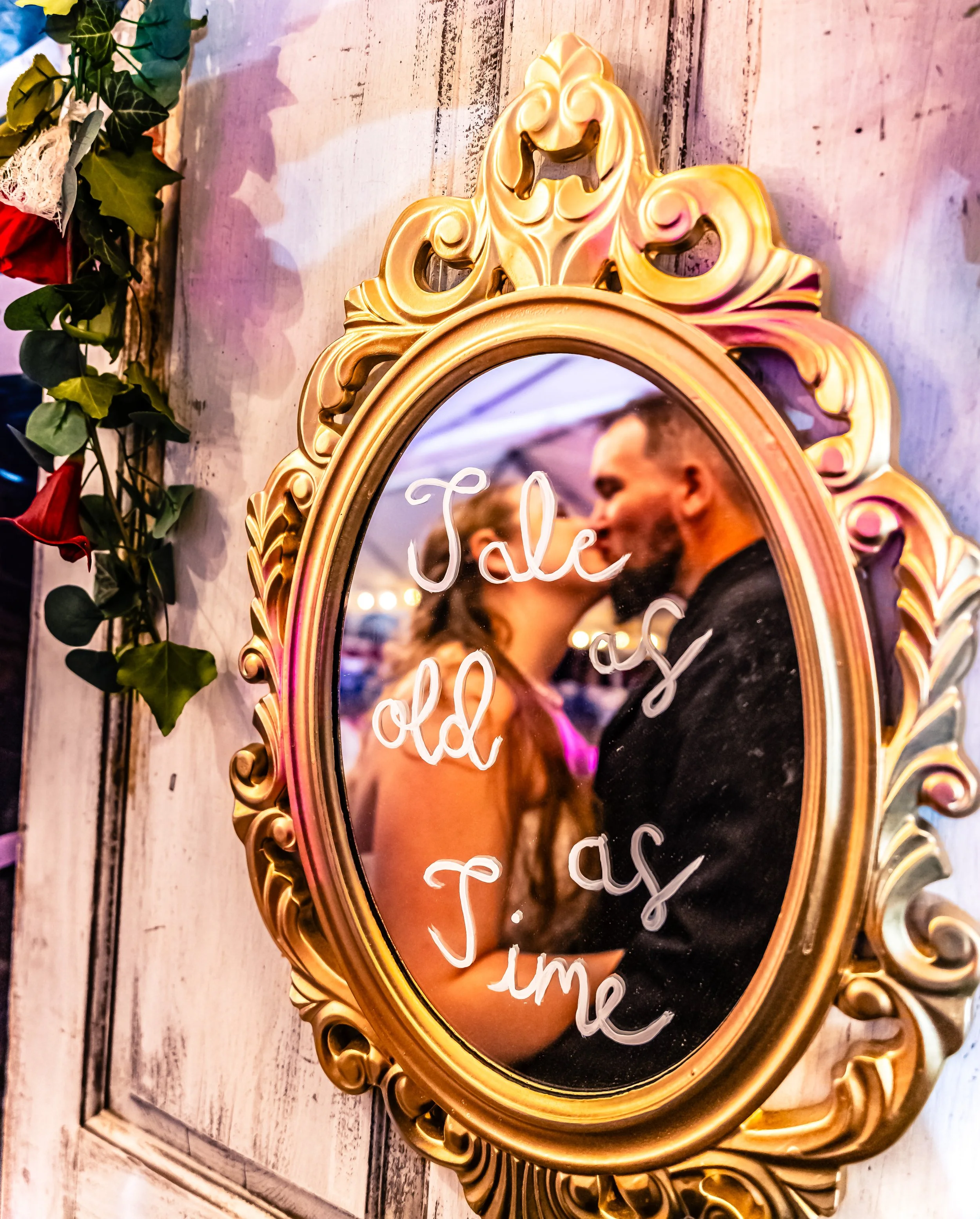 An ornate gold-framed mirror with white cursive writing that says 'Jale as old as T ime' reflecting a kissing couple at a wedding or romantic event, with a decorated wall and floral arrangement nearby.
