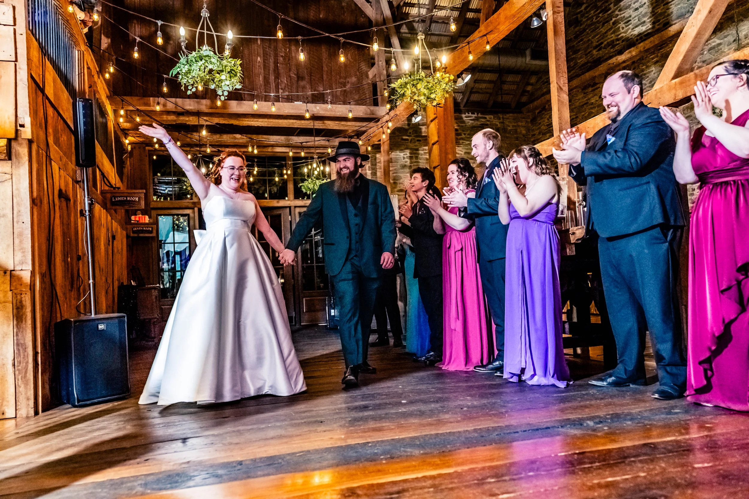 Bride and groom holding hands in the center of a celebration, surrounded by friends and family clapping and smiling in a rustic venue with hanging lights and wooden decor.