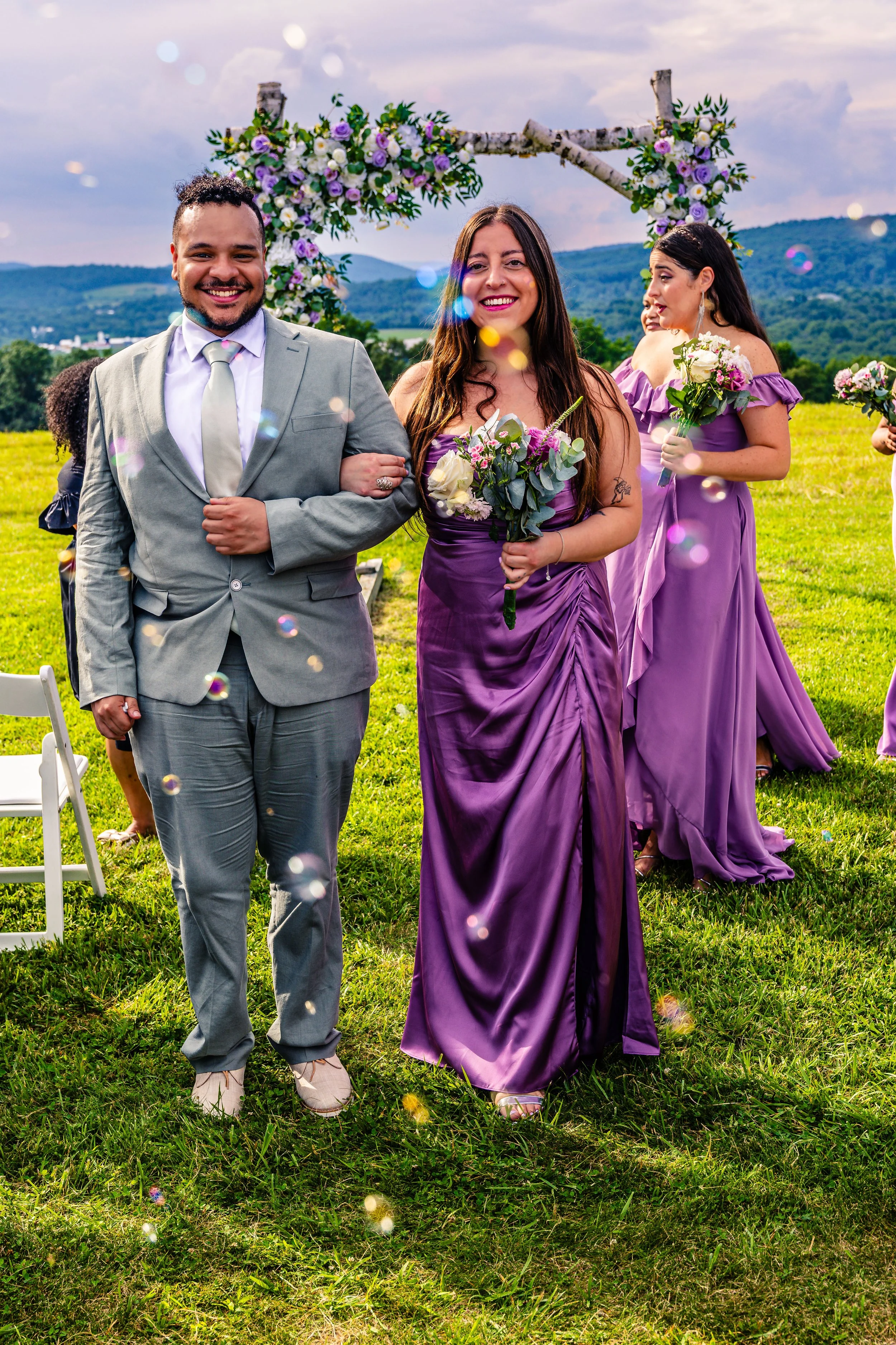 A joyful couple at an outdoor wedding, with the man in a light gray suit and the woman in a purple dress, walking arm in arm on a grassy field, with bridesmaids in matching purple dresses holding bouquets, and a scenic mountain backdrop with a floral
