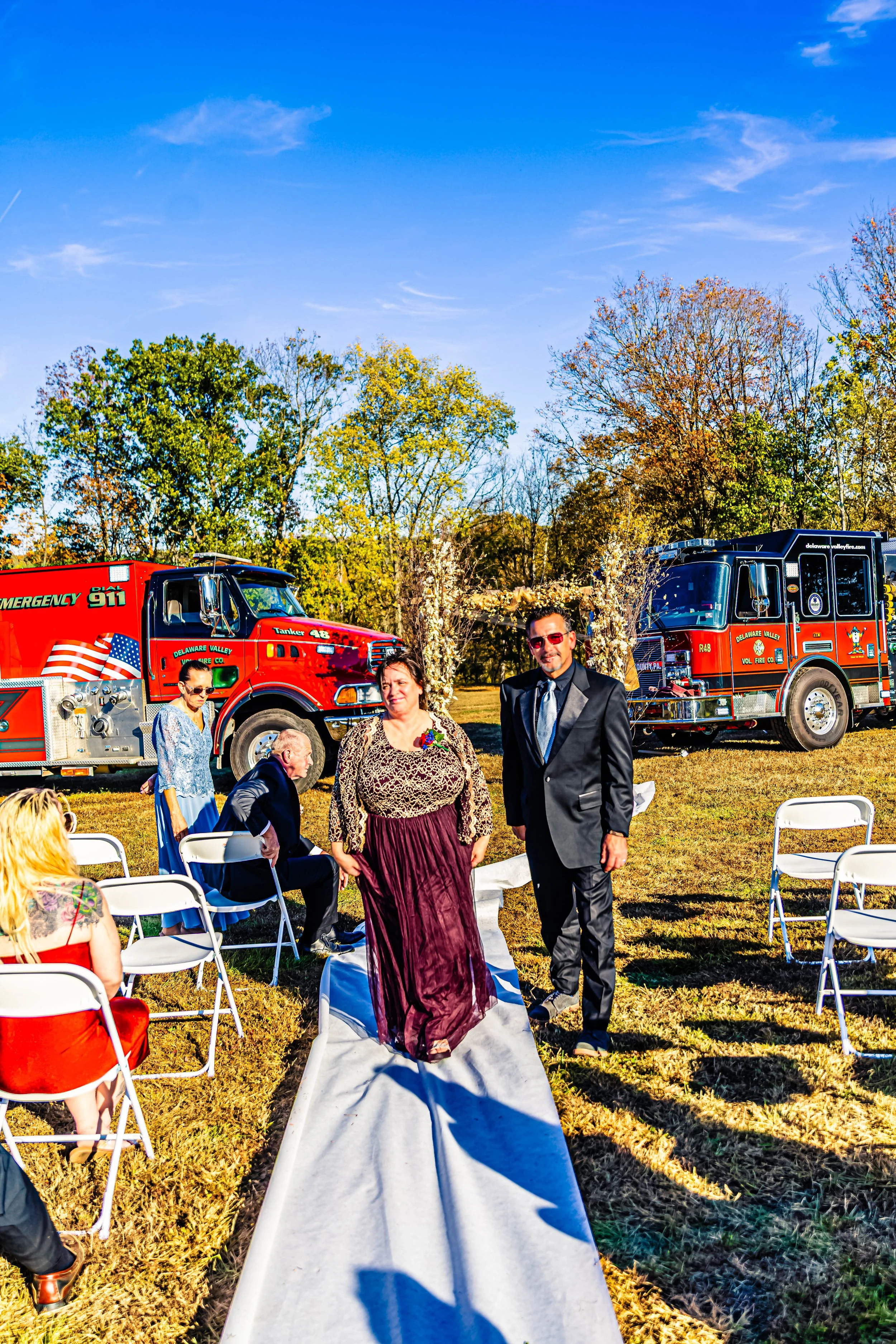 People at an outdoor wedding ceremony with two fire trucks in the background under a clear blue sky, surrounded by trees with fall foliage.