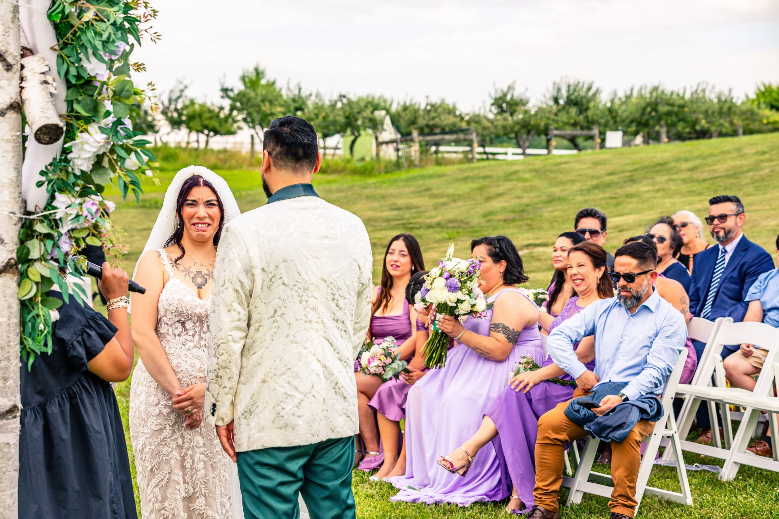 A outdoor wedding ceremony with the bride crying and the groom facing her. Guests are seated on white chairs, some smiling and holding bouquets, on a grassy field with trees and a fence in the background.