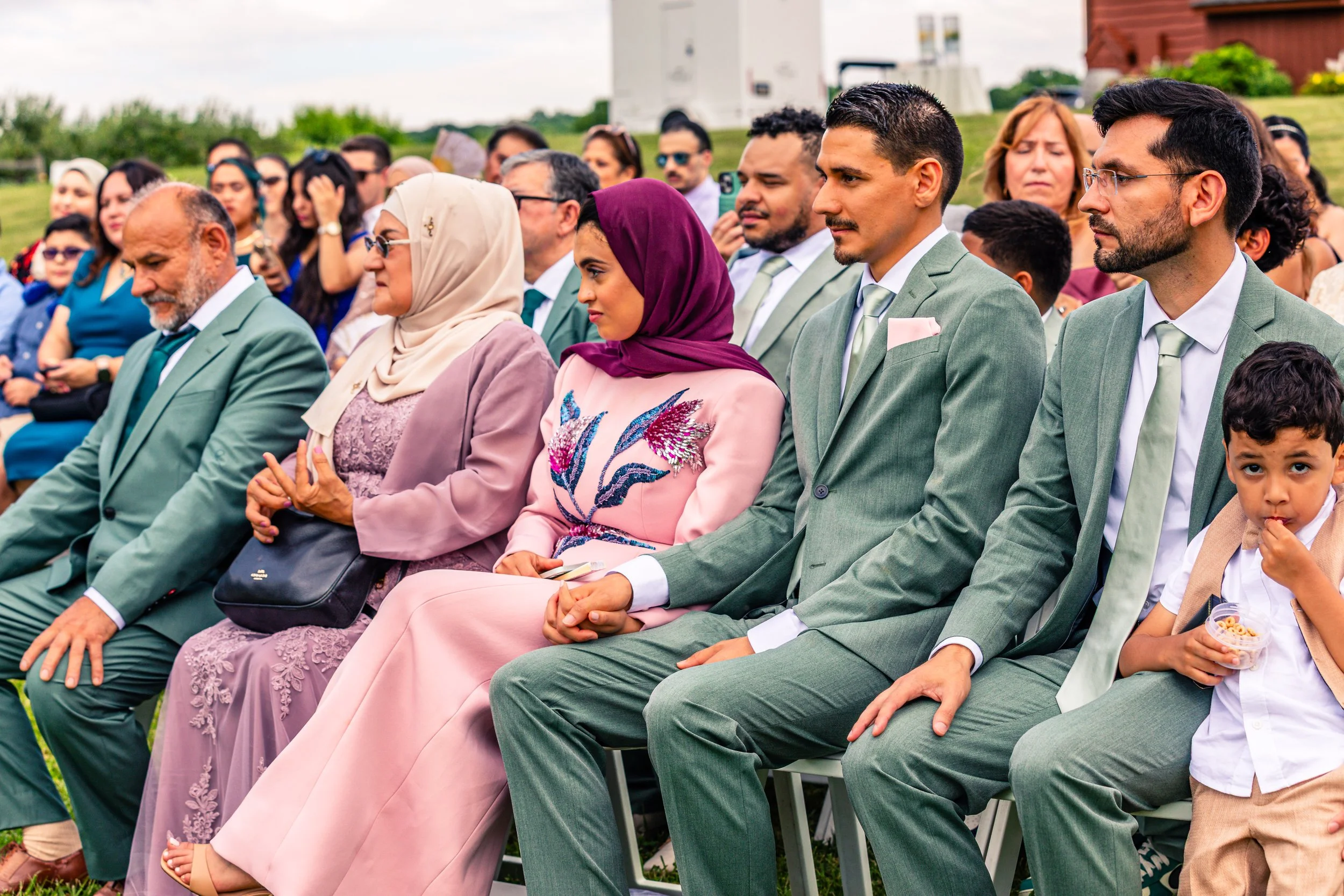 A diverse group of people dressed in formal attire, seated outdoors at a wedding or event, with some wearing traditional and contemporary clothing, and a backdrop of greenery and a cloudy sky.
