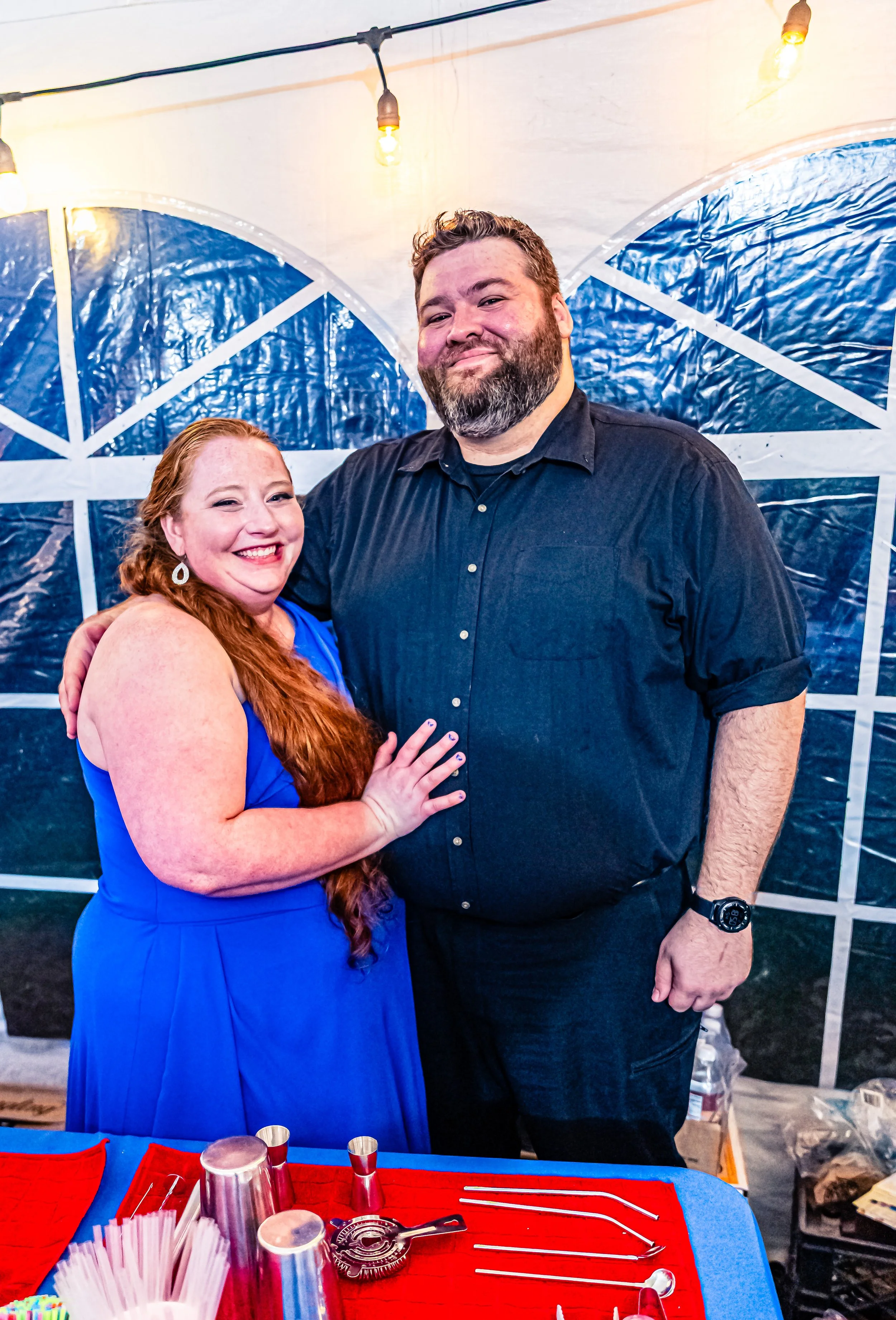 A smiling red-haired woman in a blue dress and a bearded man in a black shirt stand close together inside a white tent with string lights, posing for a photo.