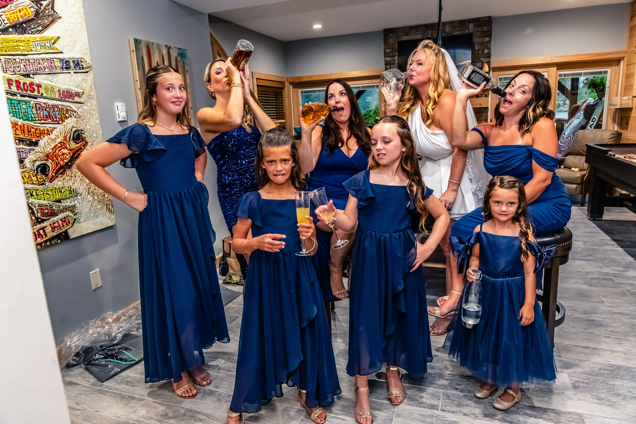 A group of women and girls wearing matching dark blue dresses in a living room, holding drinks, with some bottles raised, making celebratory gestures.