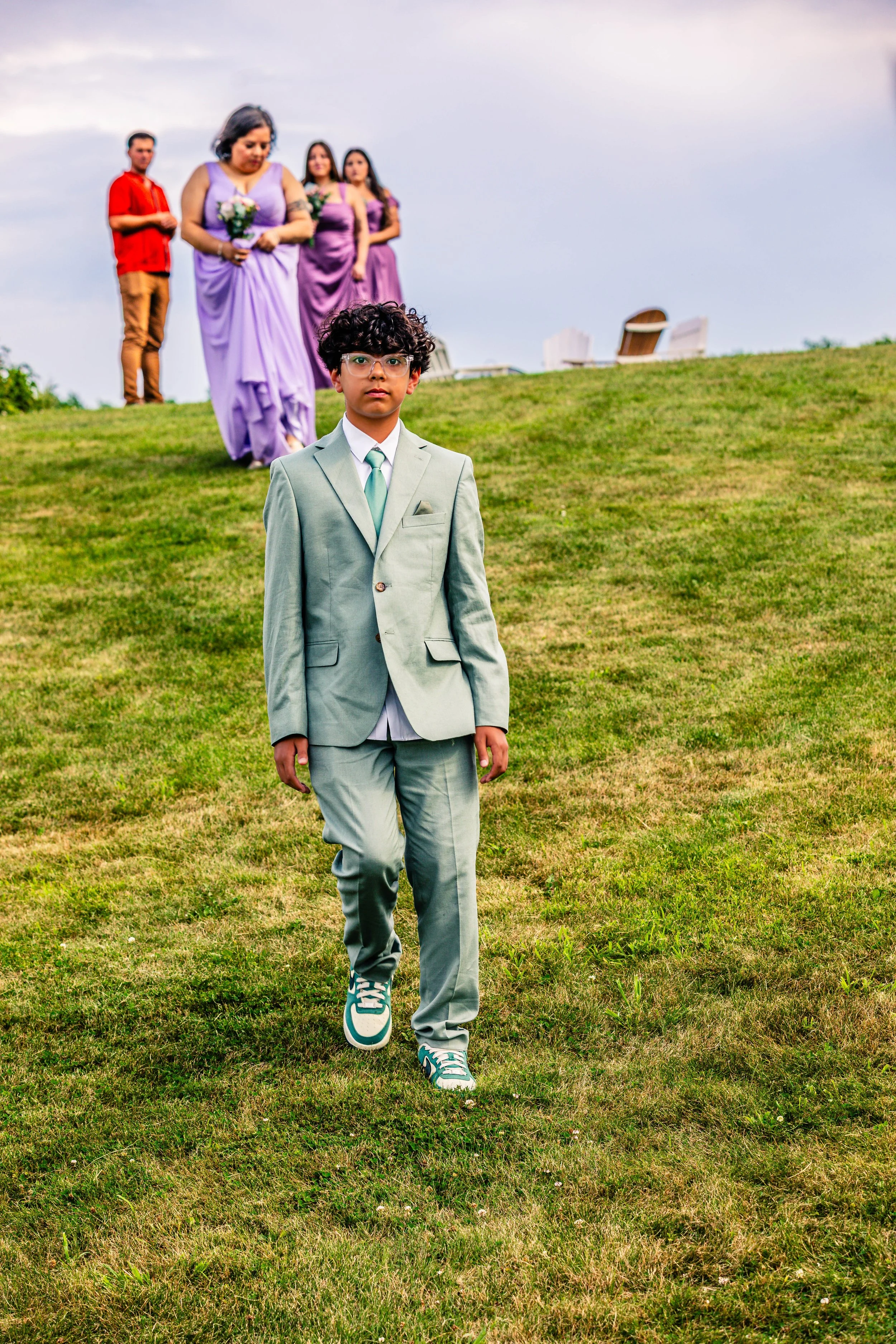 A young boy in a light gray suit and sneakers walking on a grassy hill, with four people in purple dresses and a man in red in the background, under a cloudy sky.