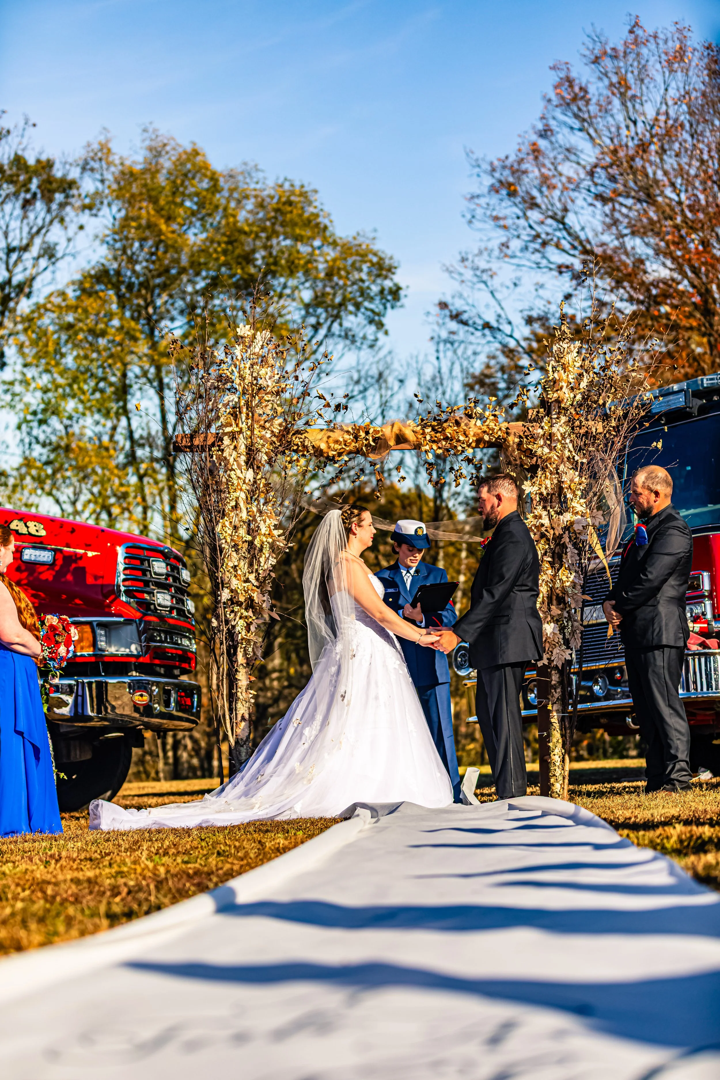 A wedding ceremony outdoors during autumn, with a bride and groom exchanging vows beneath a decorative arch made of branches and dried leaves, flanked by fire trucks and surrounded by trees with colorful fall foliage.