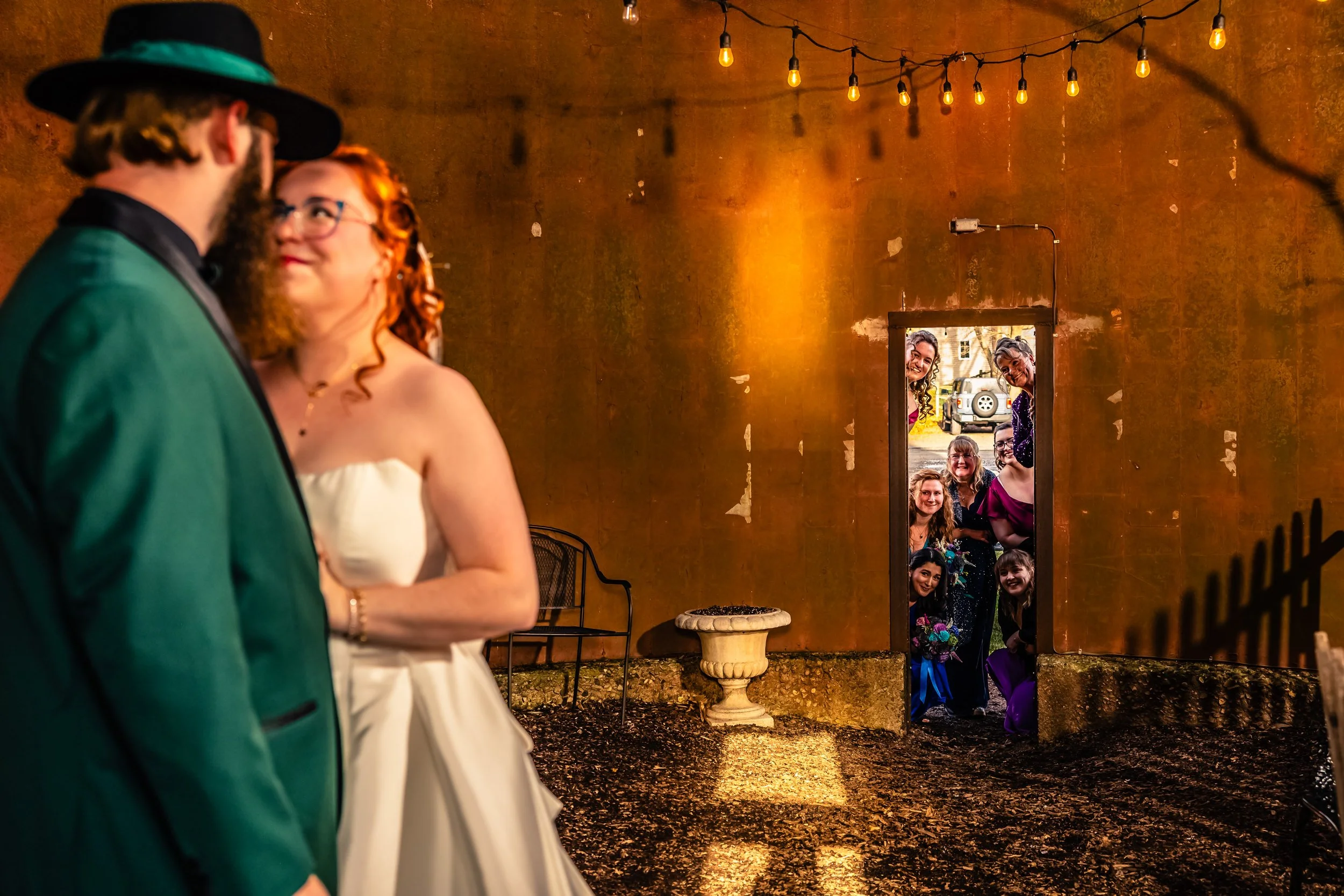 A bride and groom stand close together, facing each other, with a group of smiling women standing outside a doorway, visible through the open door, watching them. The scene is illuminated by string lights overhead.