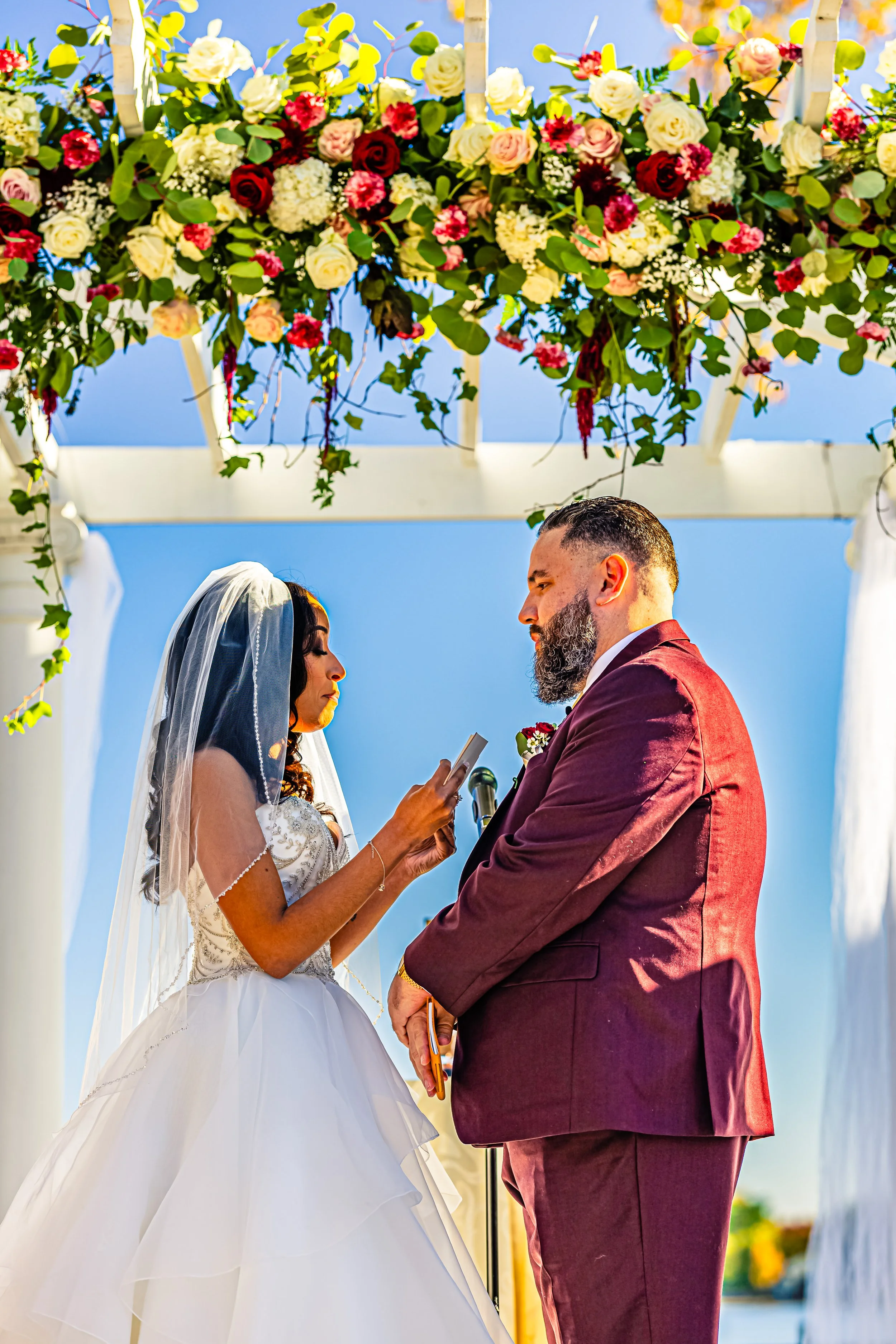 A bride and groom stand facing each other under a floral arch at an outdoor wedding ceremony. The bride is wearing a white wedding gown and veil, holding a small paper, while the groom wears a maroon suit. Bright blue sky and colorful flowers in the 