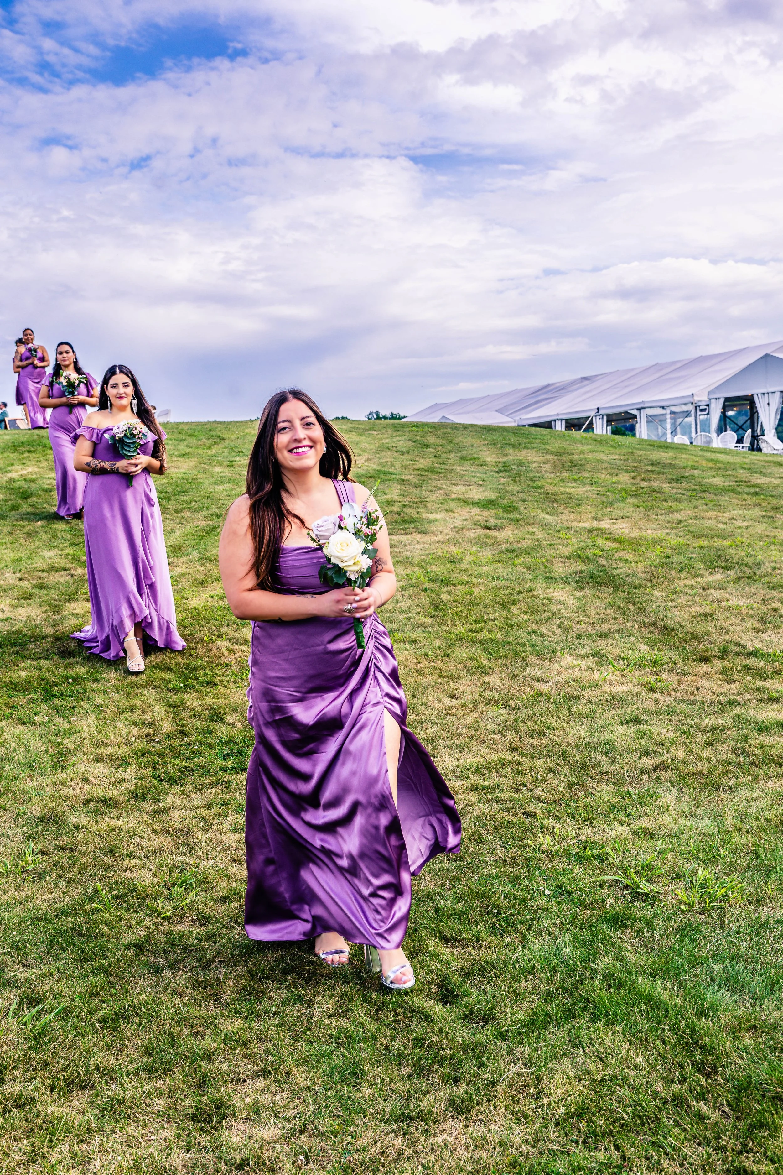 A group of women in lavender bridesmaid dresses walking down a grassy hill at an outdoor wedding, holding bouquets of white flowers, with a tent in the background under a cloudy sky.