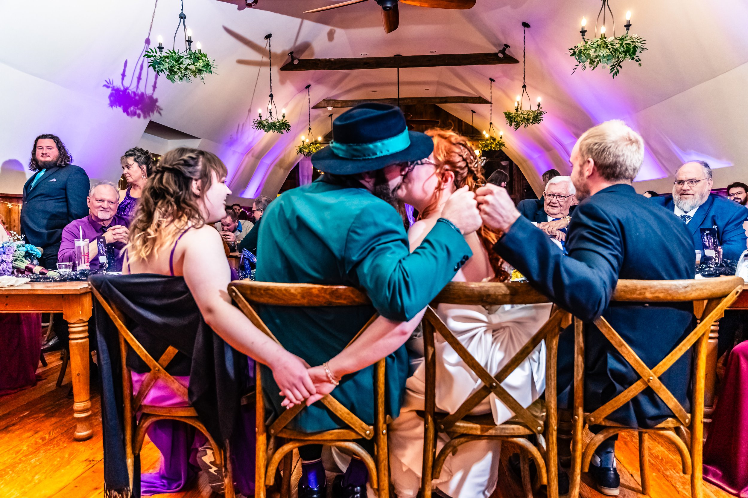 A couple kisses at a wedding reception surrounded by guests, including a girl in a purple dress and older people, with hanging chandeliers and floral decorations in the background.