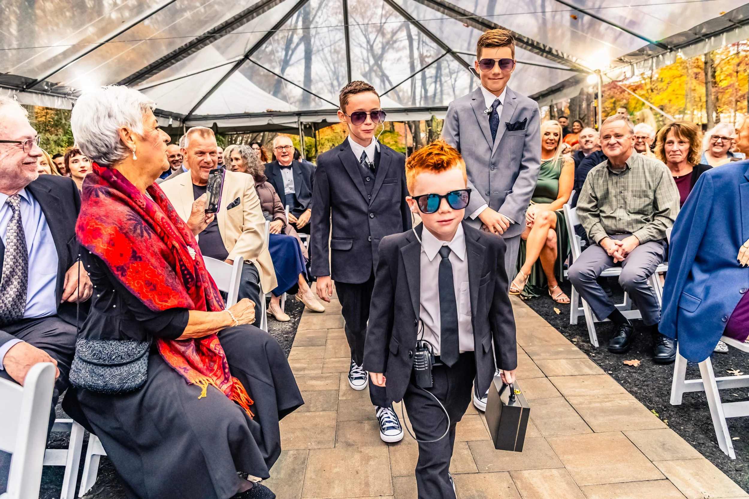 Four young boys in suits and sunglasses walking down an aisle at an outdoor wedding or formal event, with seated guests watching under a canopy in autumn.