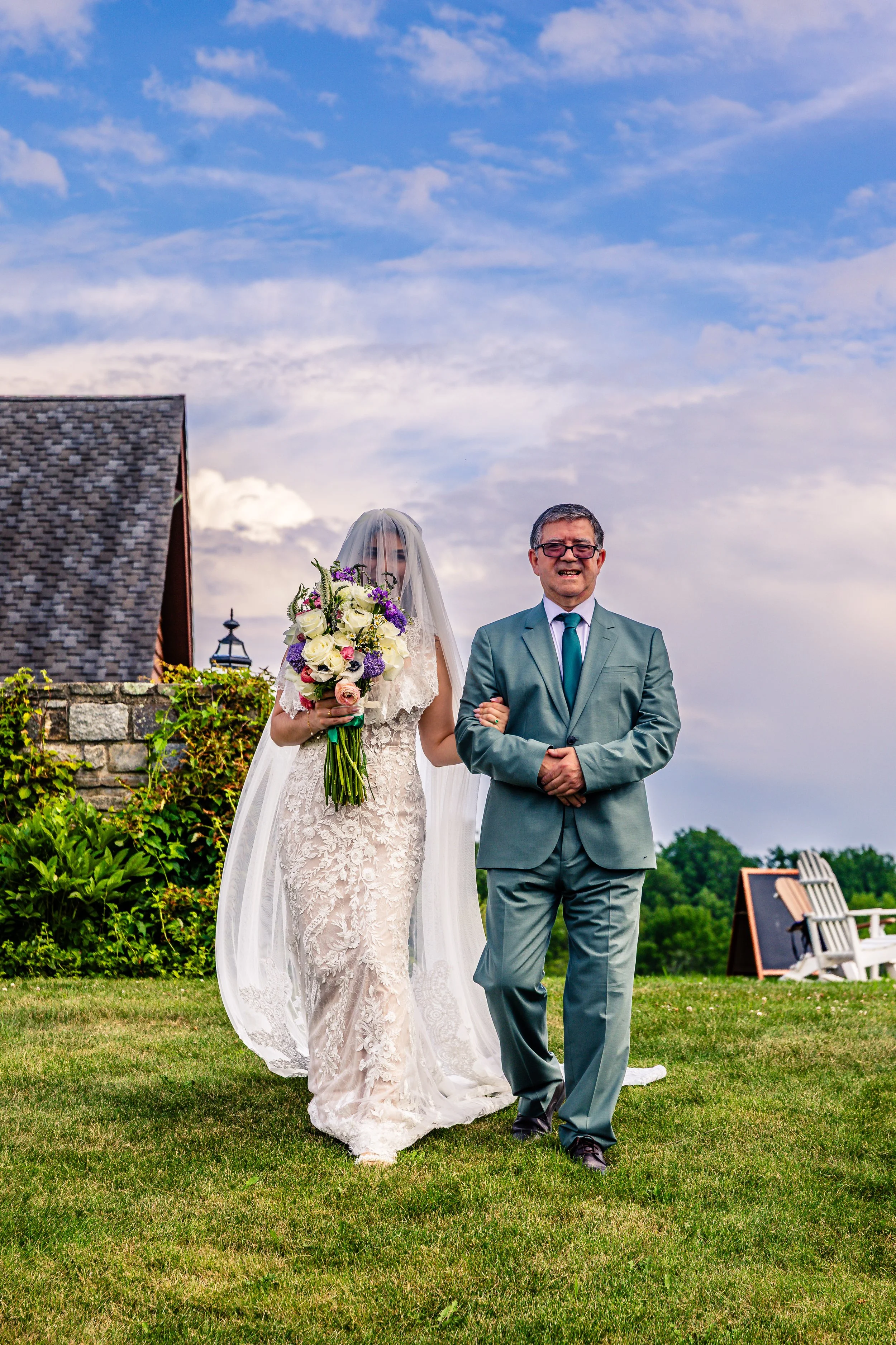 A bride in a lace wedding gown holding a bouquet of flowers walking down the aisle with an older man, possibly her father, during an outdoor wedding ceremony.
