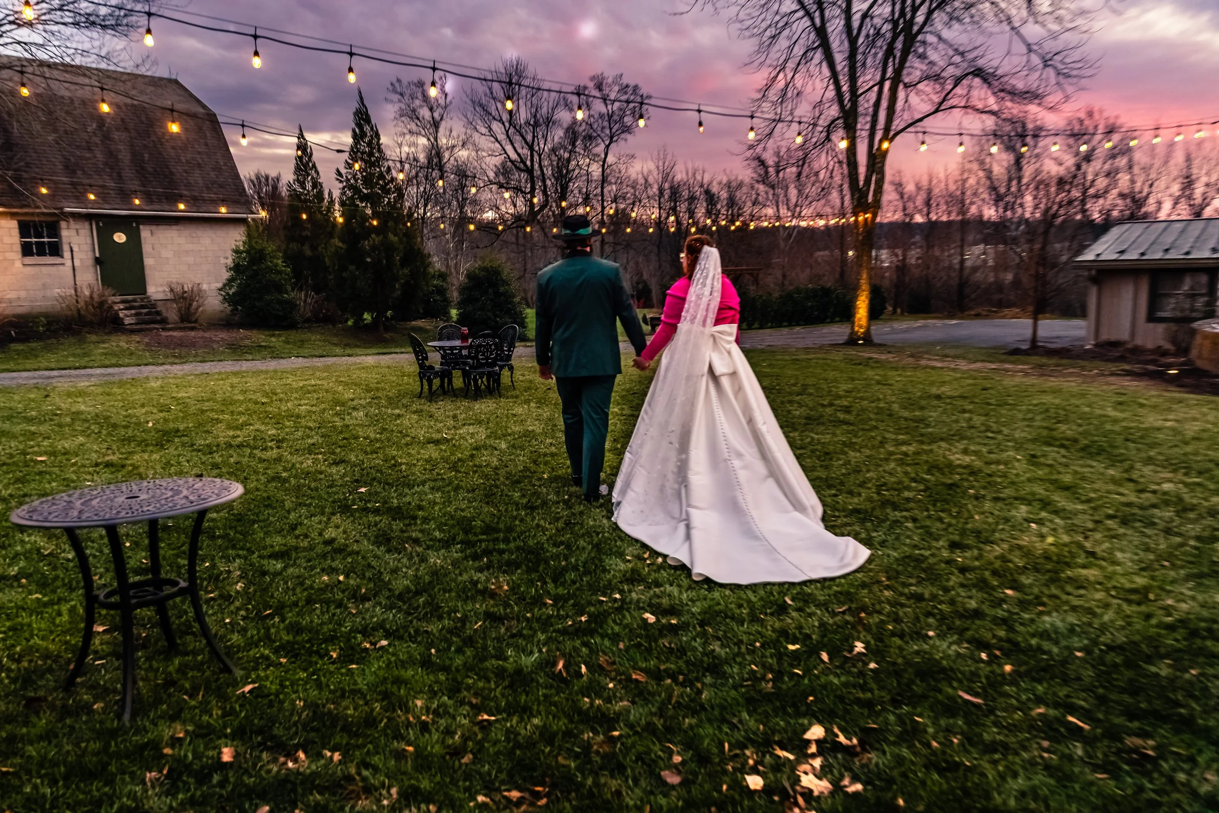 A bride and groom holding hands walking in a garden at sunset, with string lights hanging overhead and trees in the background.