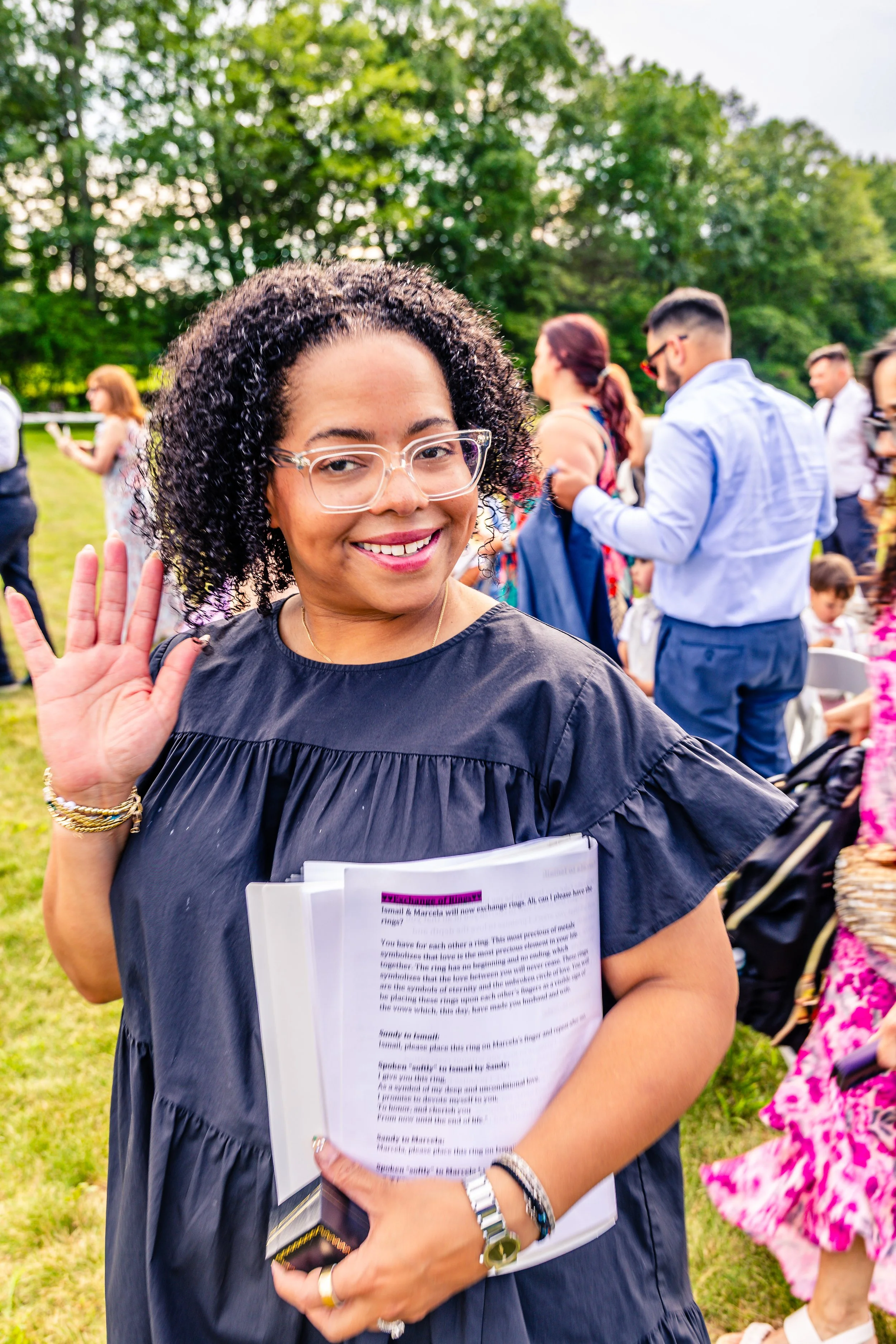 Smiling woman waving at outdoor event, holding papers, wearing glasses and a black dress, with seated and standing people in the background.