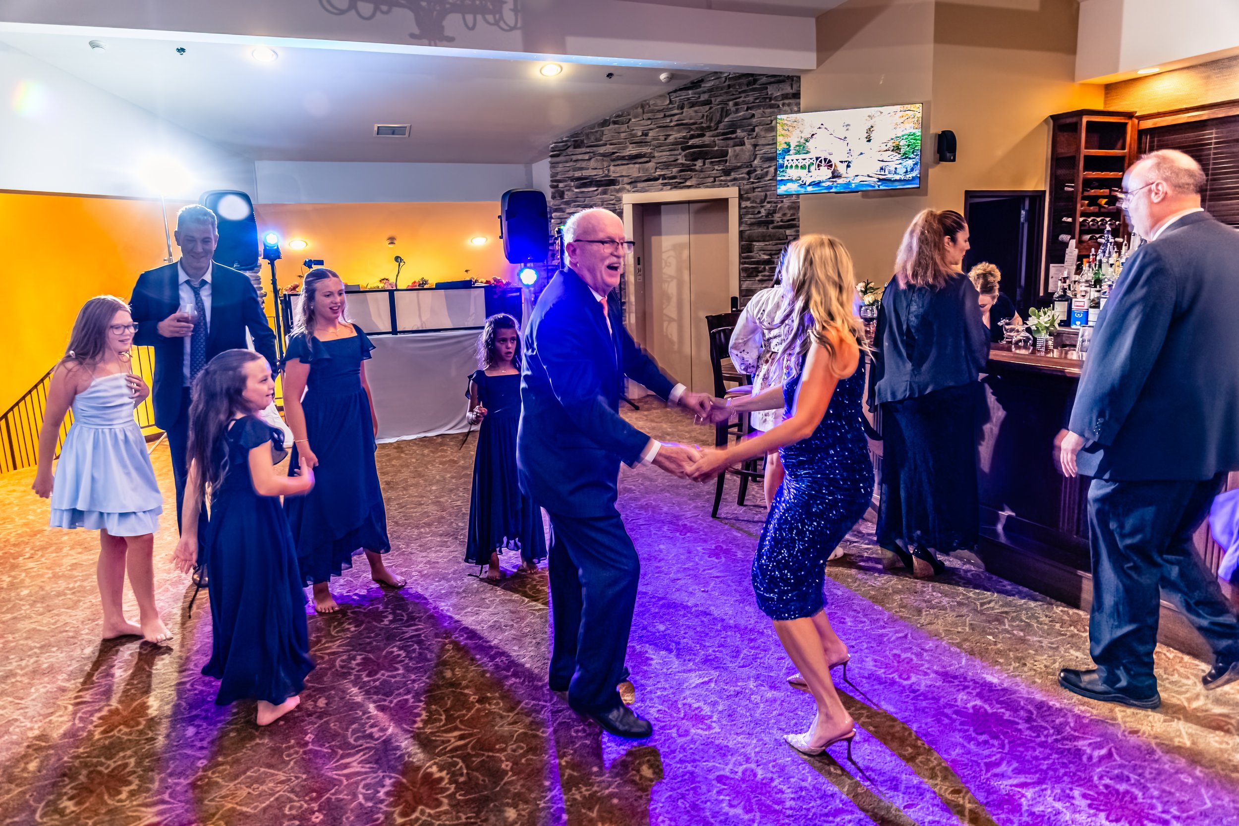People dancing and enjoying themselves at a party in a decorated indoor venue with a bar, barstools, and a stone fireplace in the background.