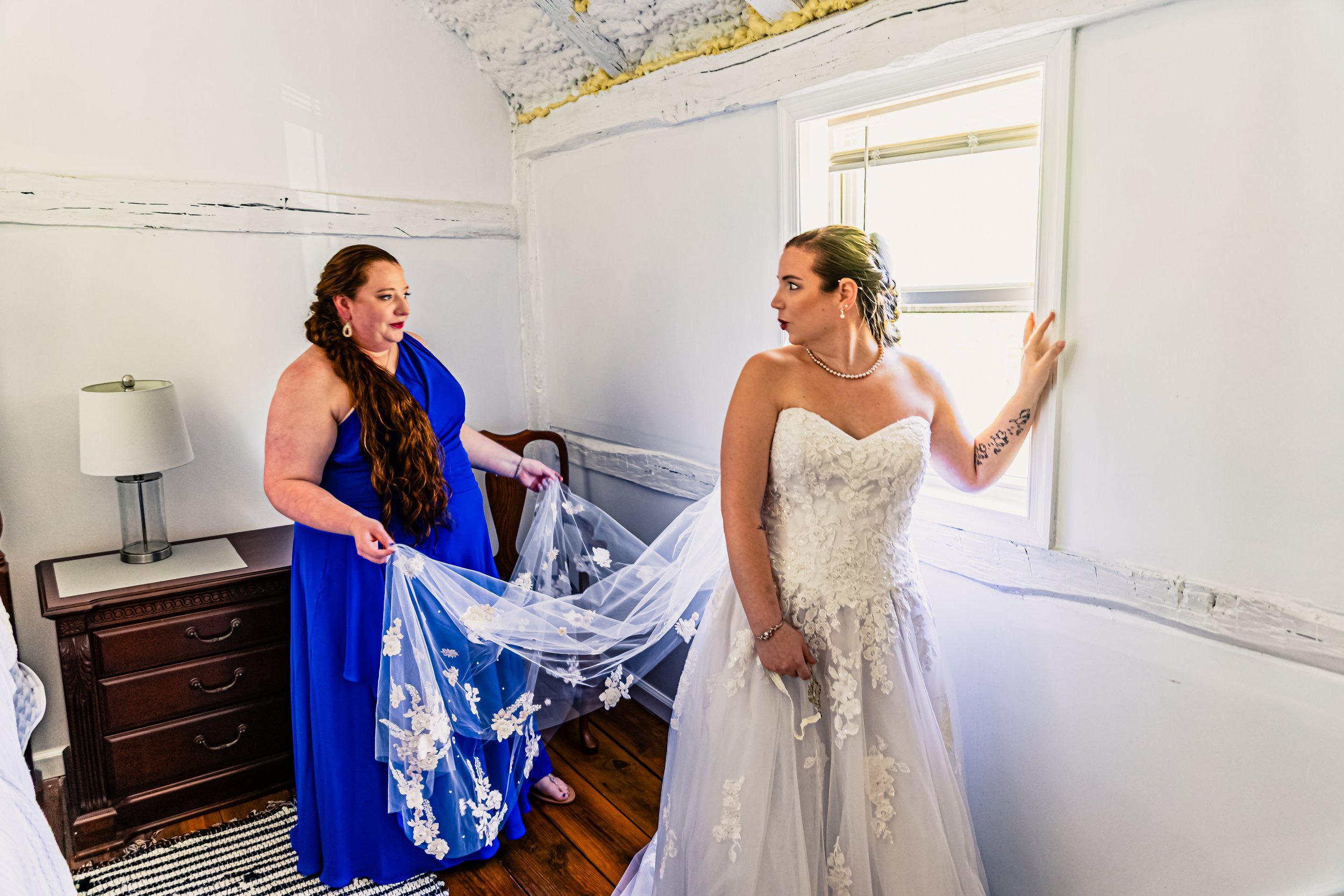 A bride in a white lace wedding dress standing by a window, with a bridesmaid in a blue dress holding her train in a small, bright room.