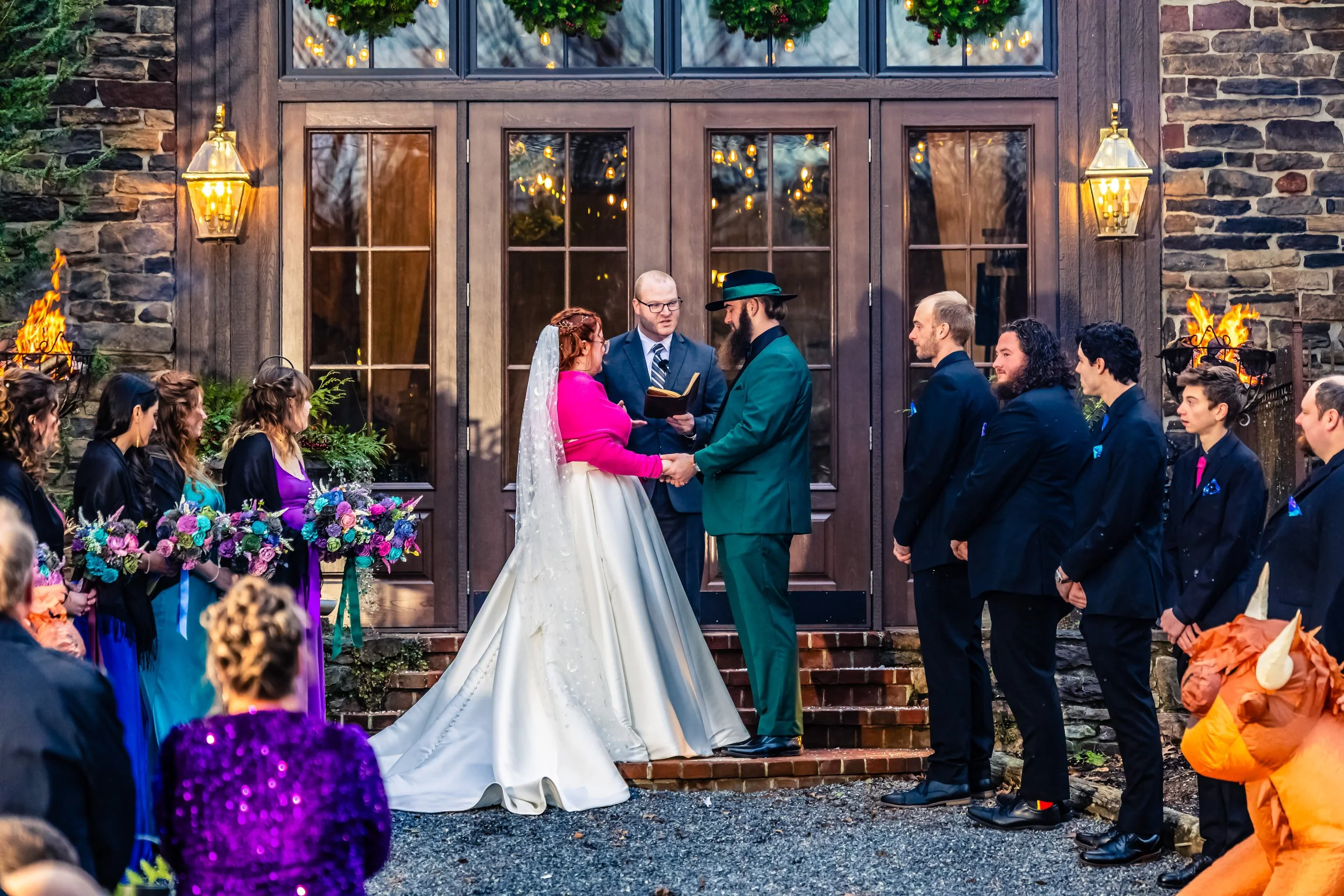 A wedding ceremony taking place outdoors in front of a rustic building with large glass doors. The bride and groom are holding hands and facing each other, with a person officiating in between. The bride is wearing a white dress with a pink shawl and