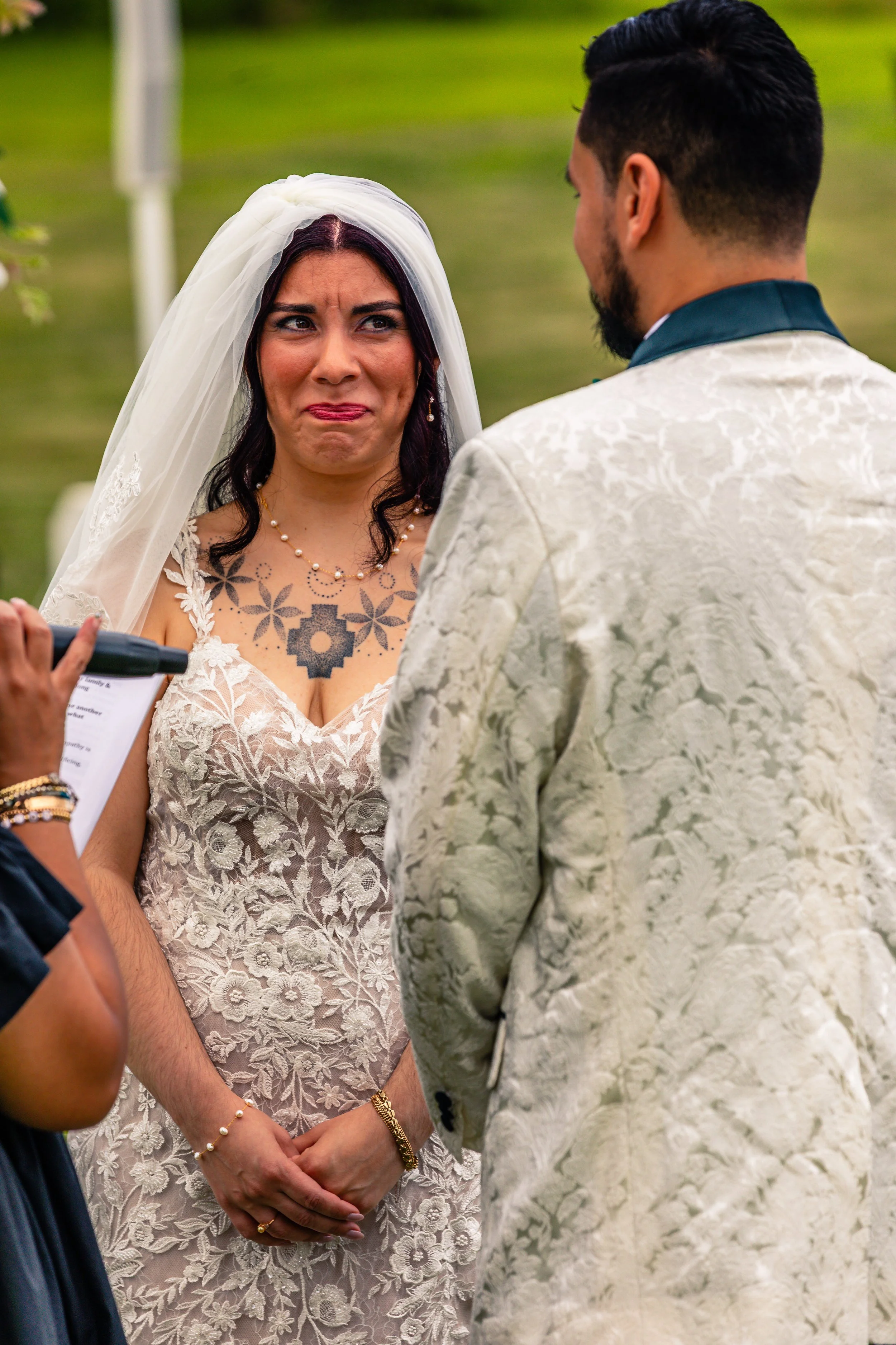 A woman with a tattoo on her chest looking emotional during a wedding ceremony, facing a man in a cream-colored tuxedo with a floral pattern, outdoors with green grass in the background.